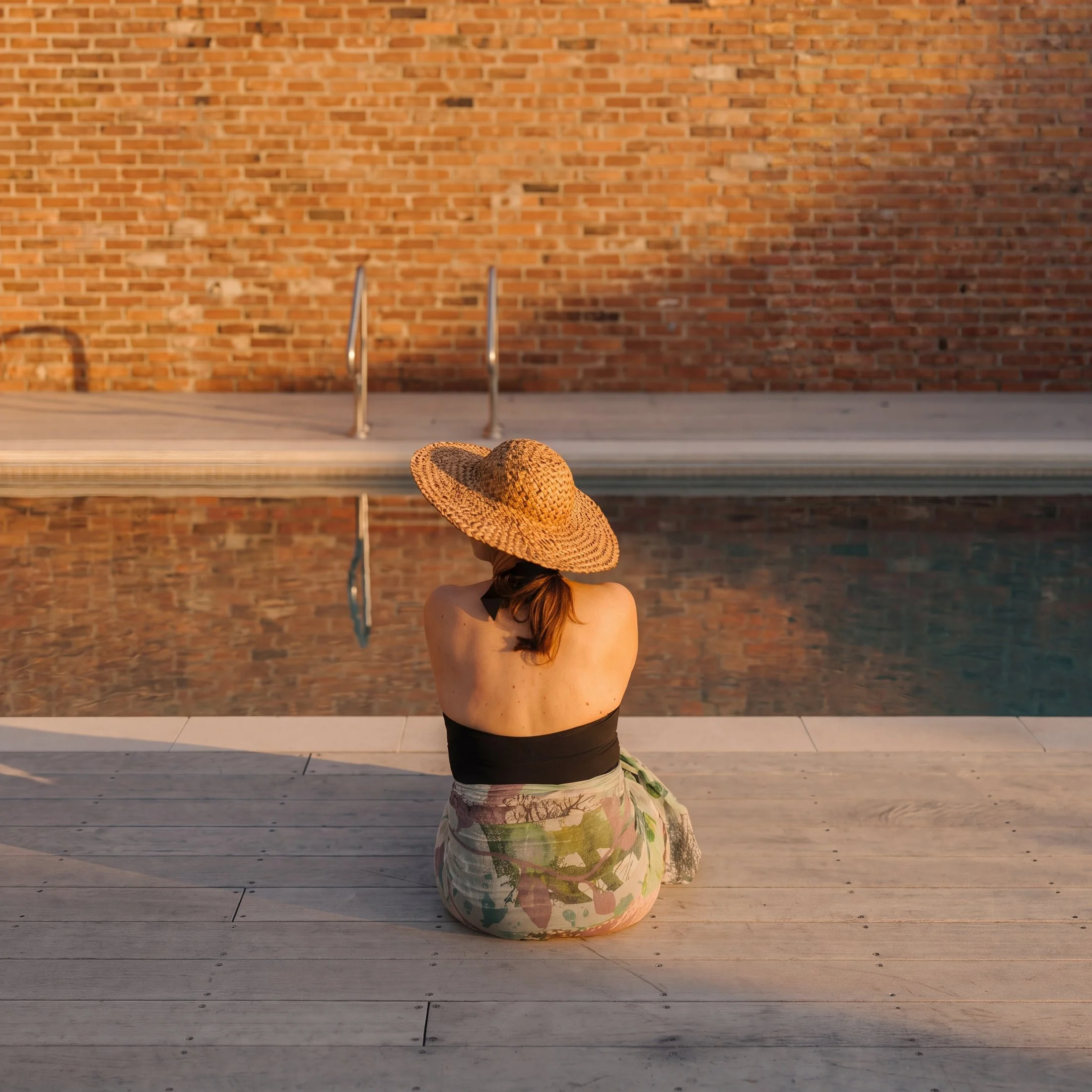 A woman, sitting by a pool, wearing a sun hat and a floral sarong, facing away from the camera towards a brick wall.