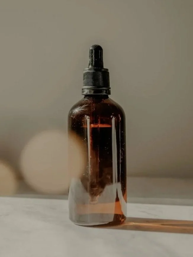 A small amber glass spray bottle with a black dropper cap, placed on a white surface, with a blurred neutral background.