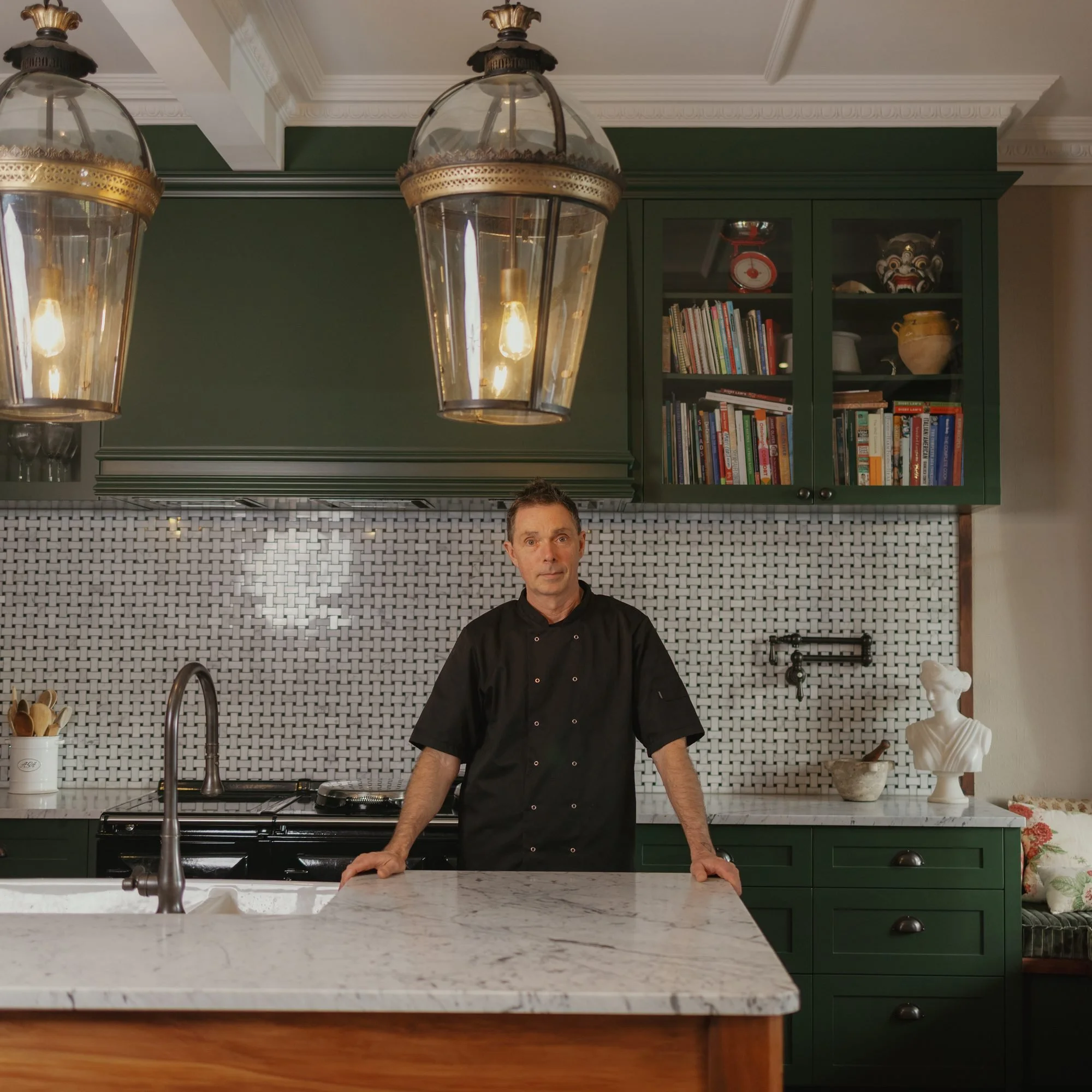 A man in a black chef coat stands behind a white kitchen island with a marble countertop, in a kitchen with dark green cabinetry, a white backsplash with a woven pattern, and decorative items including a sculpture and a bowl.