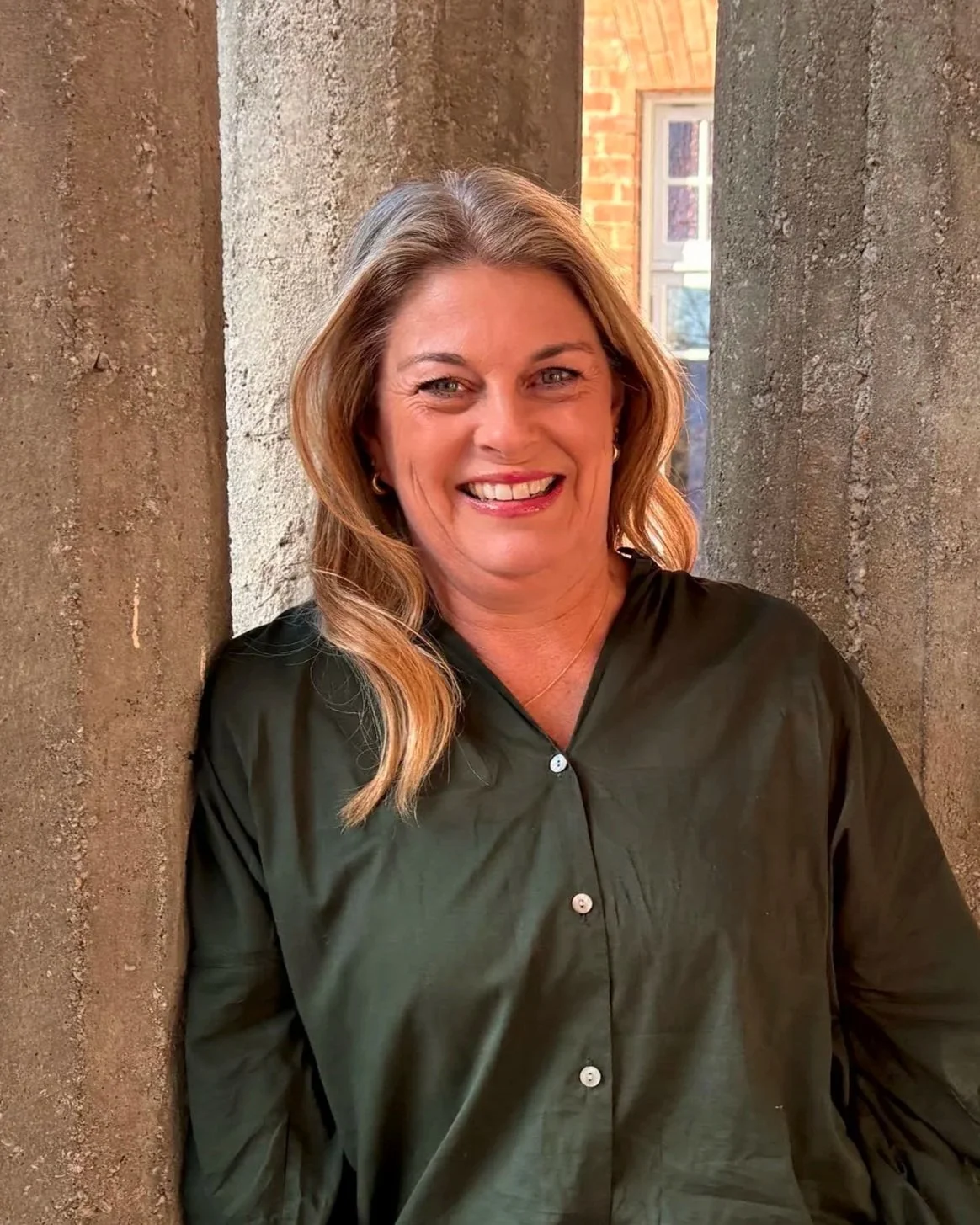 A smiling woman with wavy blonde hair, wearing a dark green button-up shirt, standing between two textured concrete pillars with a brick building and a window in the background.