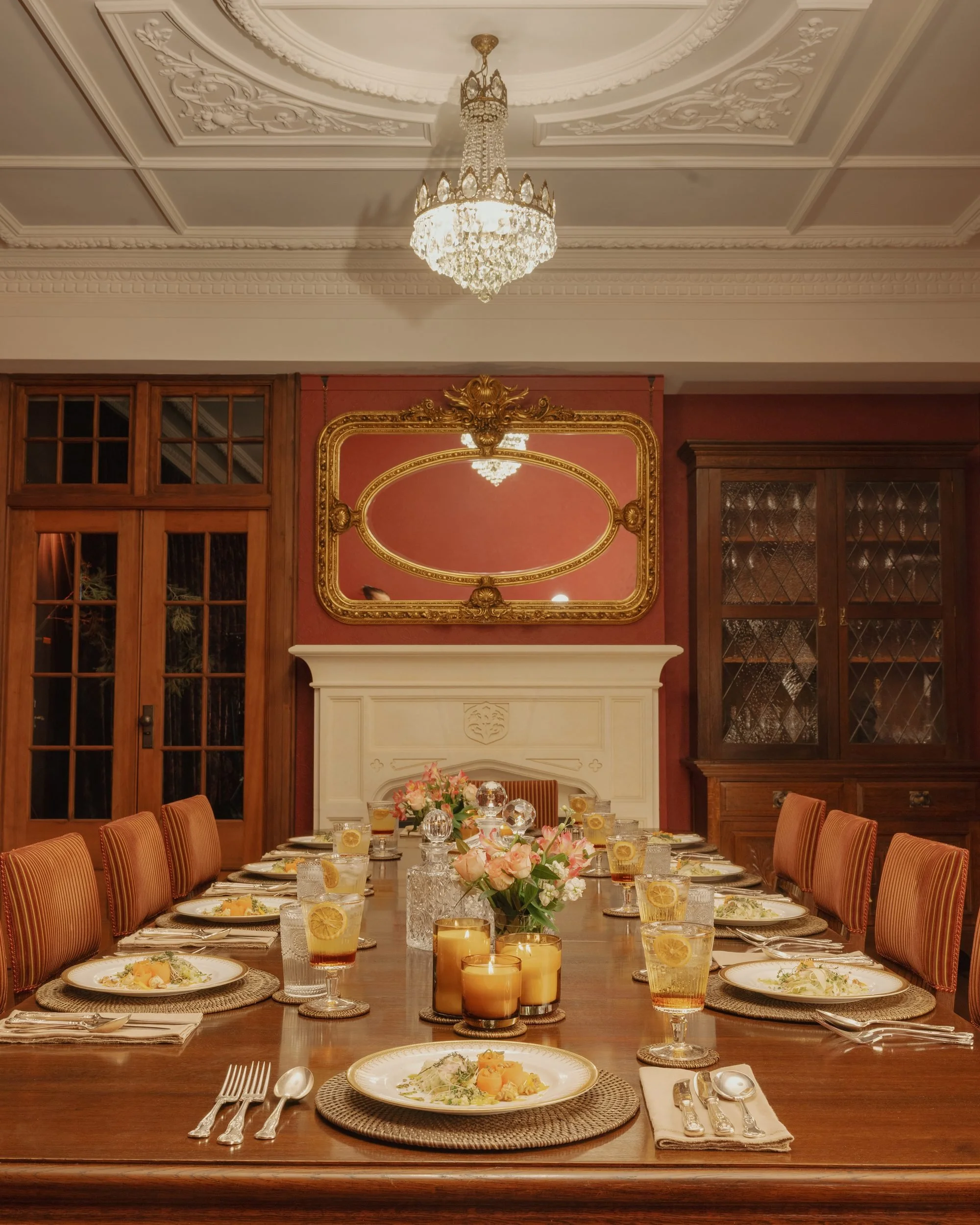 Elegant dining room with a long wooden table set for a meal, decorated with floral arrangements and candles, surrounded by upholstered chairs, with ornate wall moldings, a large mirror with gold details, and a chandelier hanging from a decorated ceiling.