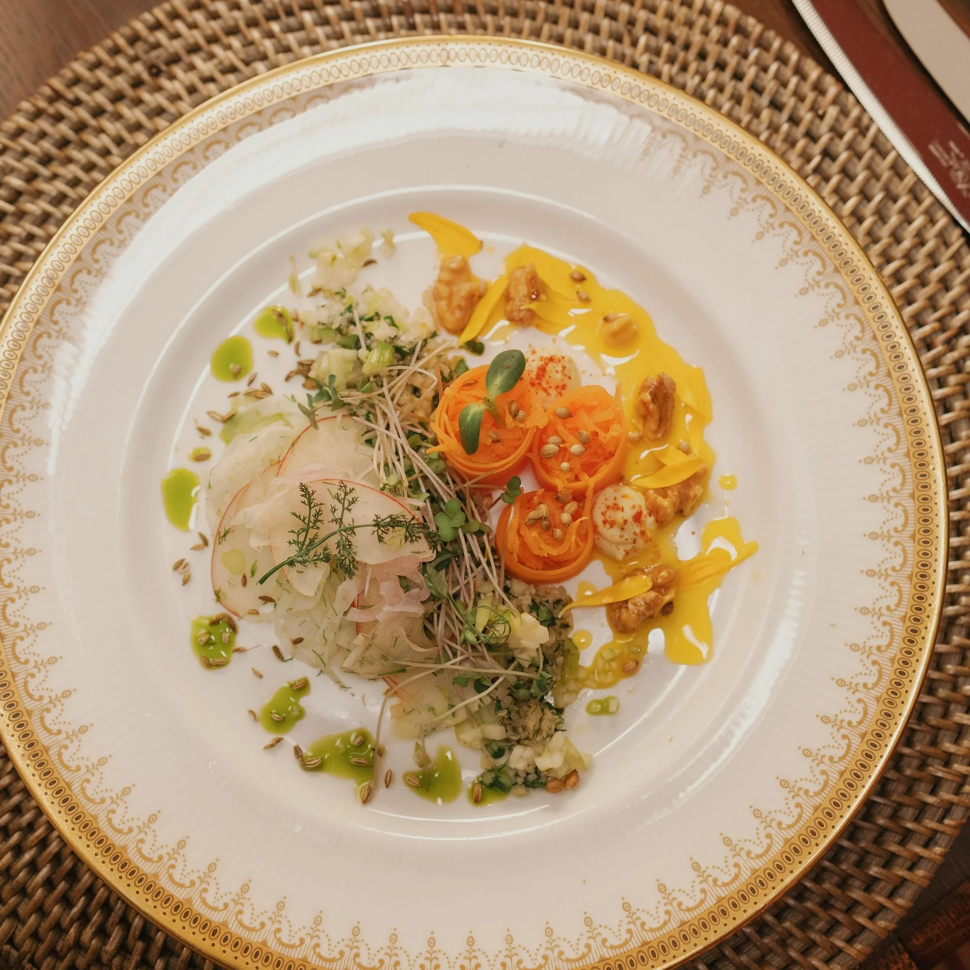 Close-up of a decorative white and gold plate on a woven placemat, with a variety of colorful food garnishes.