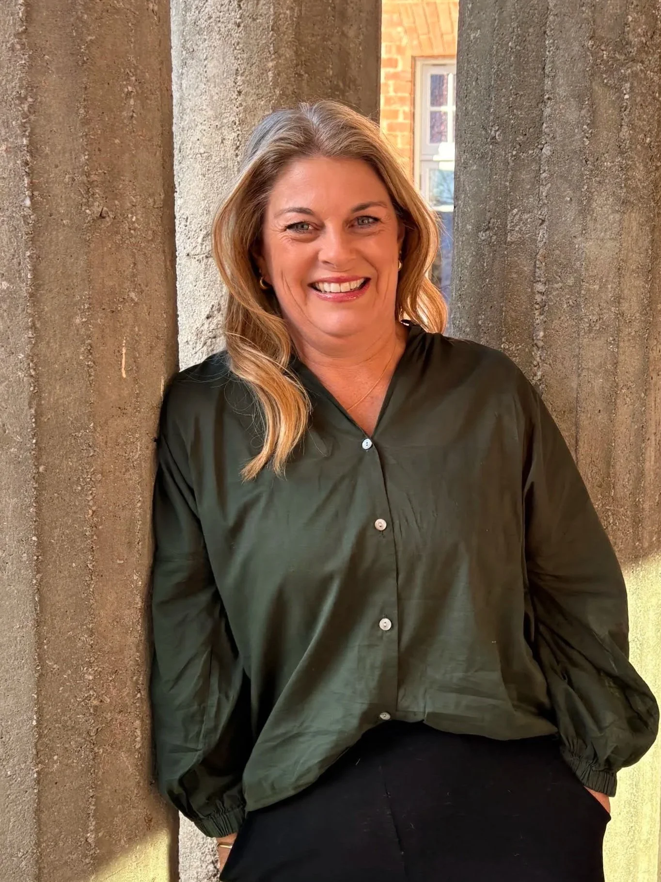 A woman with blonde hair, wearing a black shirt, standing against a textured wall with windows in the background, smiling at the camera.