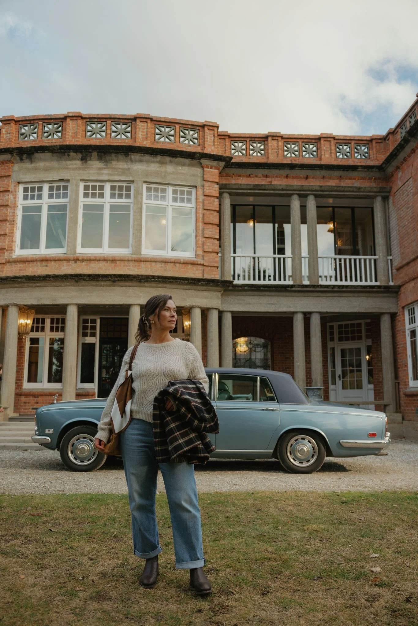 A woman wearing a beige sweater, blue jeans, and black boots, holding a plaid jacket and a tan handbag, standing in front of a vintage blue car parked outside a large brick house with columns and a balcony.