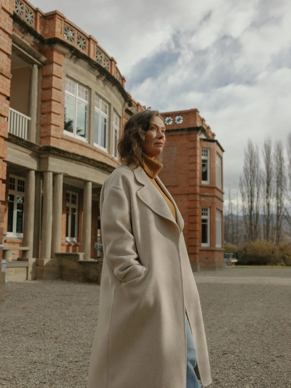 Woman in a beige coat standing outside a large brick building with columns and windows, under a cloudy sky.