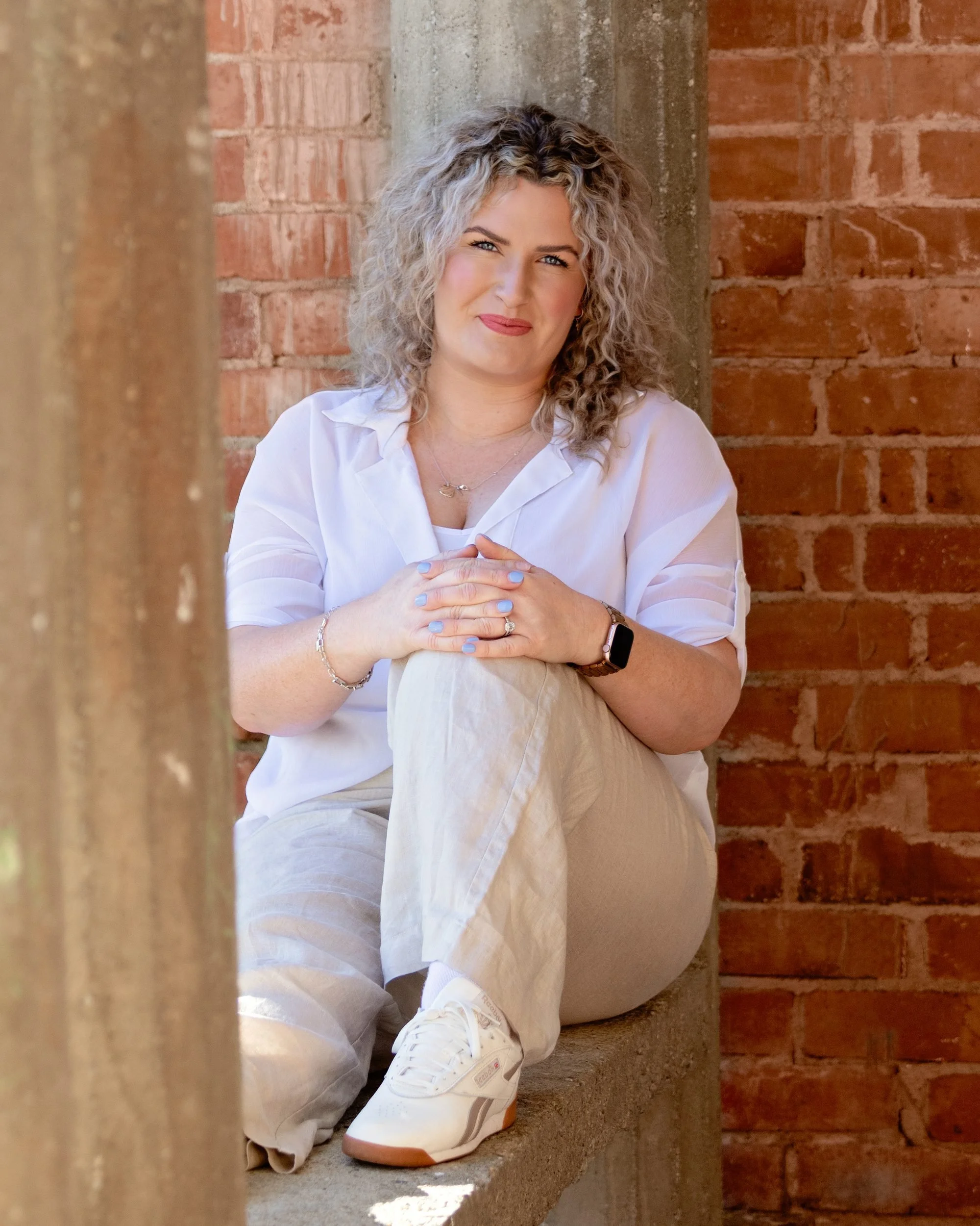 A woman with curly blonde hair sitting on a concrete ledge against an exposed brick wall. She is wearing a white shirt, light-colored pants, and white sneakers. She has her hands clasped together and is smiling slightly while looking at the camera.