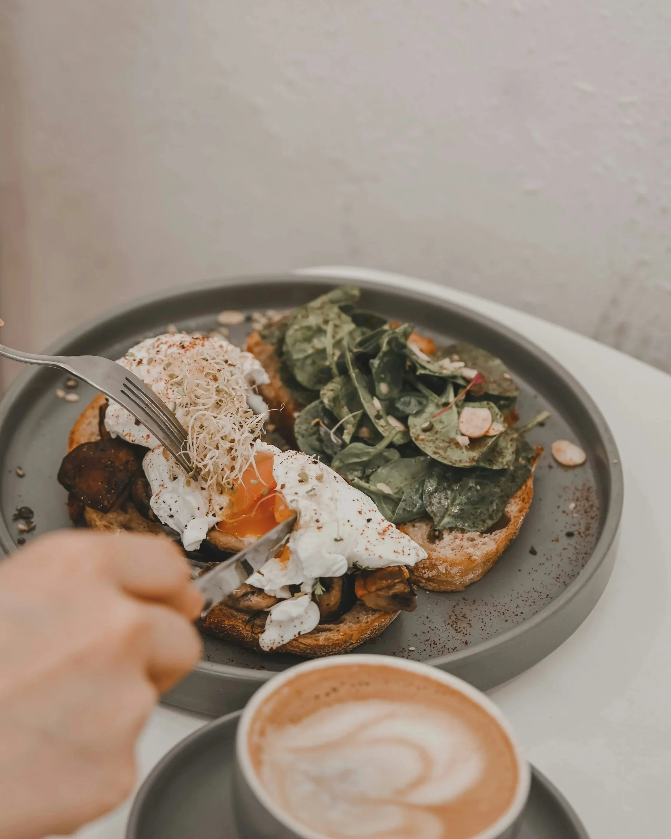 Close-up of a plate with fried egg, leafy greens, and other toppings, with a fork in hand, and a cup of latte or cappuccino in the foreground.