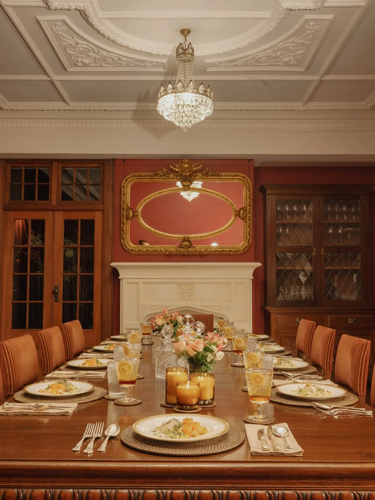 Elegant dining room with a long wooden table set for a meal, decorated with candles and floral arrangements, ornate ceiling with chandelier, and classic wood-paneled walls with cabinetry.
