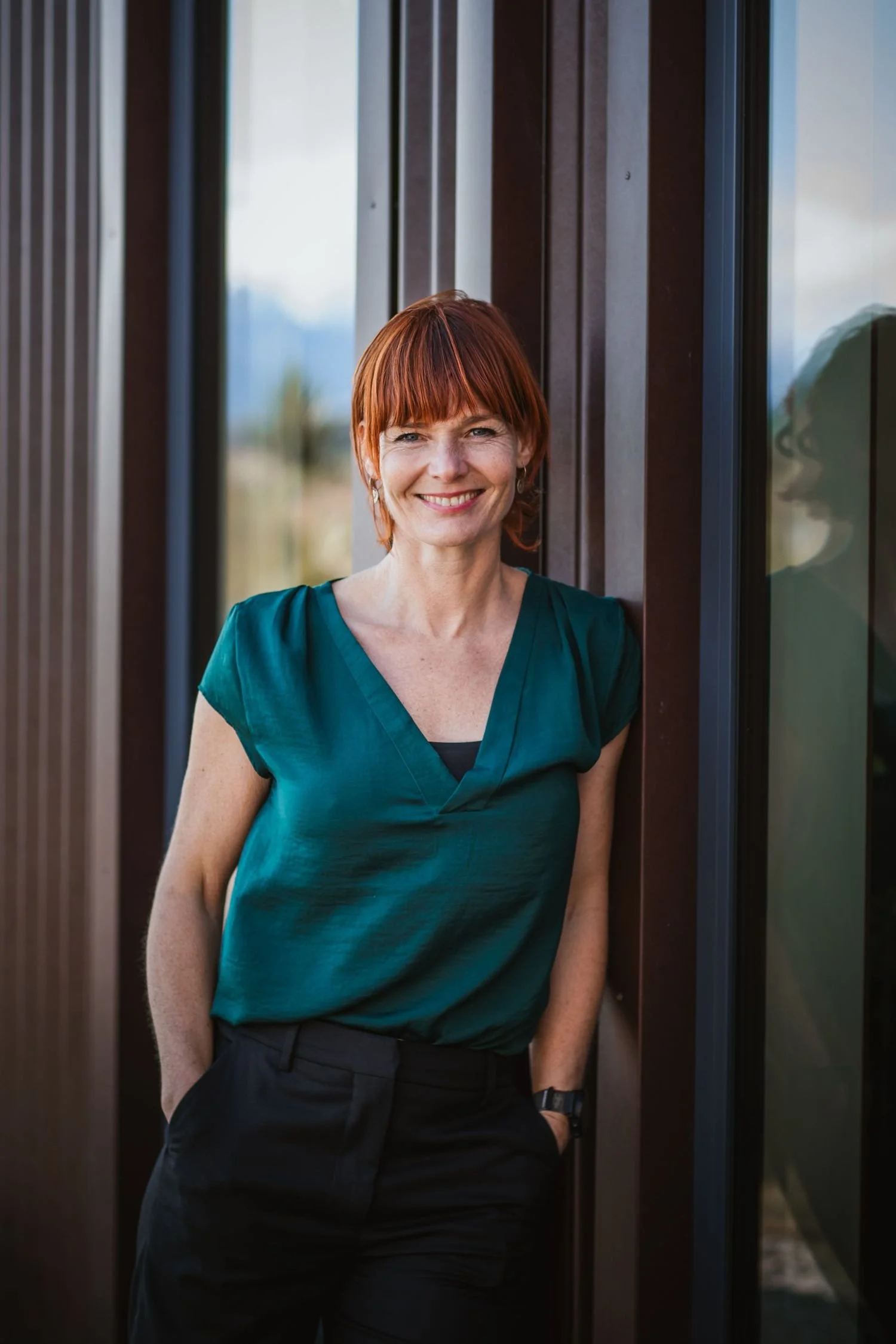 A woman with short red hair and wearing a teal shirt standing outside next to a modern building with dark brown panels and large glass windows, smiling at the camera.