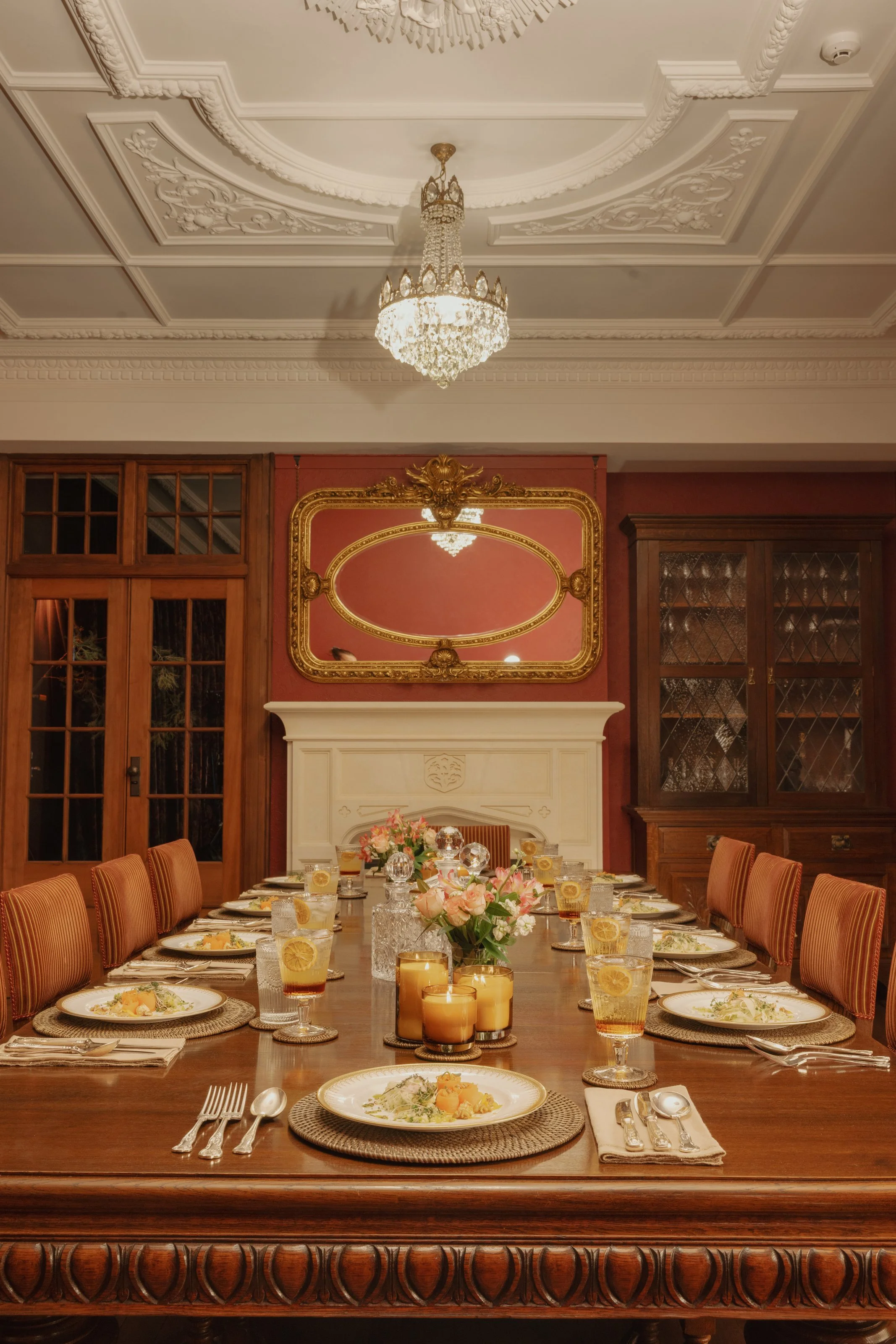 Elegant dining room with a long wooden table set for a meal, decorated with candles and floral arrangements, ornate ceiling with chandelier, and classic wood-paneled walls with cabinetry.