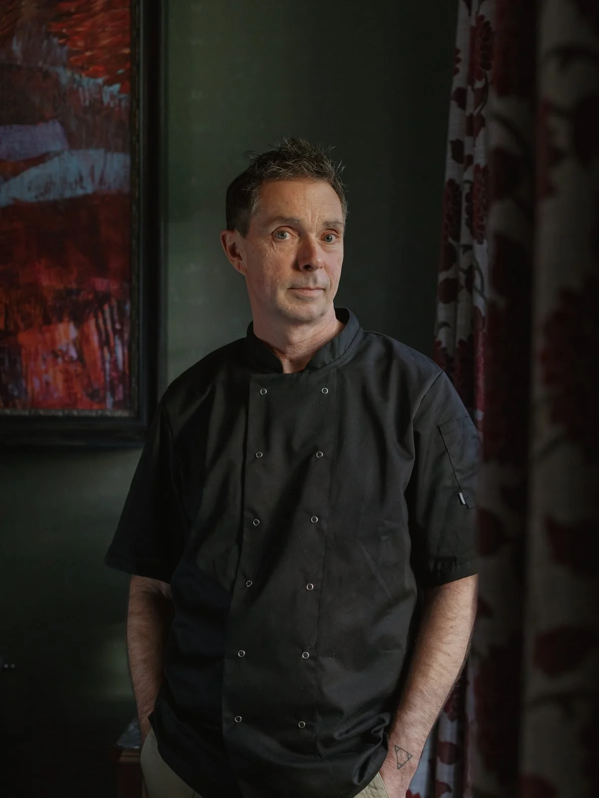 A man in a black chef's jacket standing indoors near a window with dark green walls and red-patterned curtains, looking at the camera.