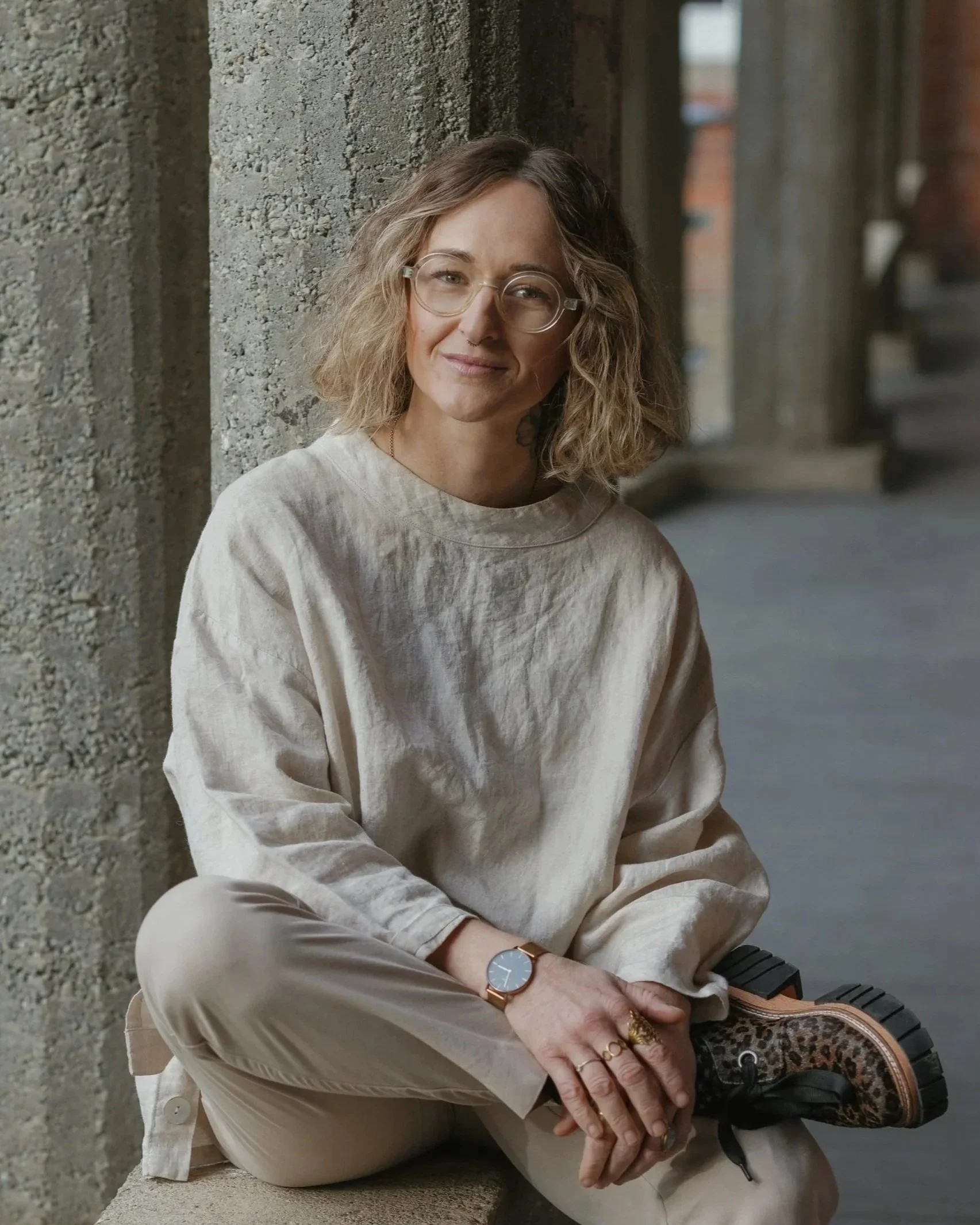 A woman with curly blonde hair and glasses sitting on a bench next to a concrete wall, holding her hands in her lap, wearing a beige sweater and a watch.