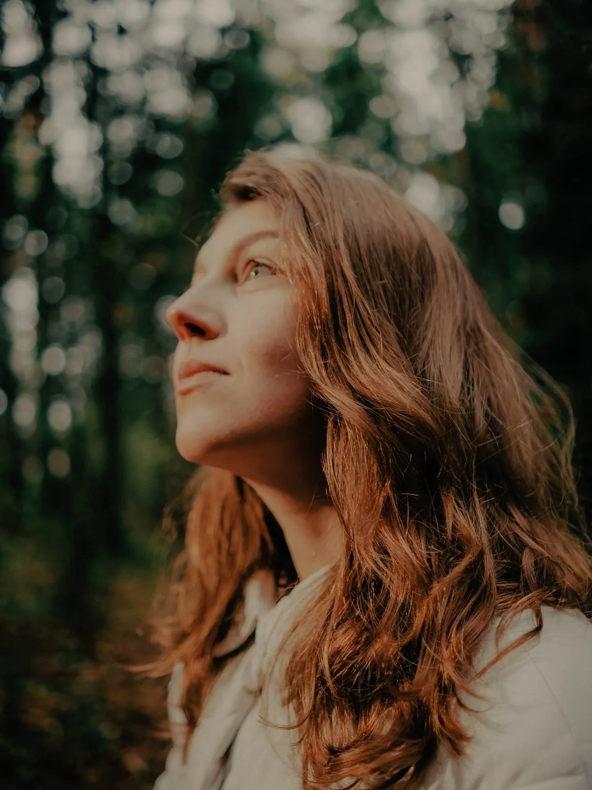 A woman with long, reddish-brown hair gazing upwards outdoors with a blurred background of trees and foliage.