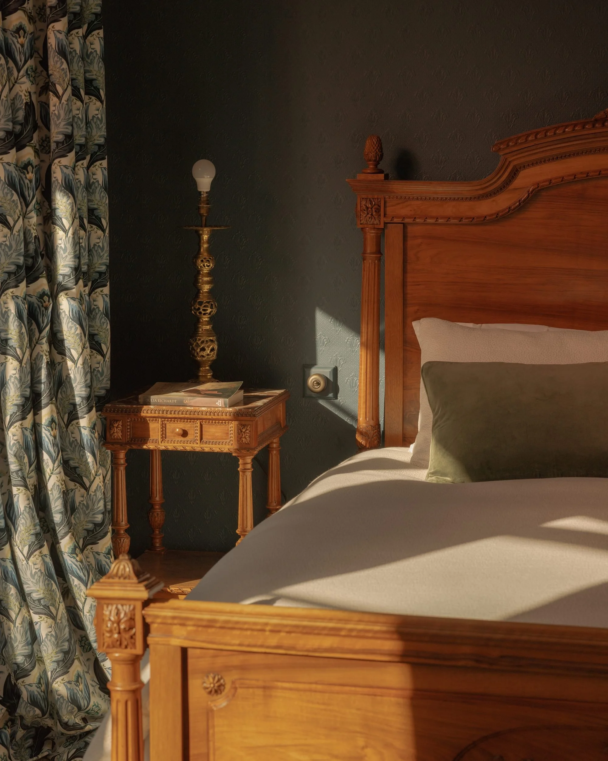 A bedroom corner with a wooden bed frame, a nightstand with books, a vintage brass floor lamp with a white bulb, dark patterned wallpaper, and a curtain with a floral design.