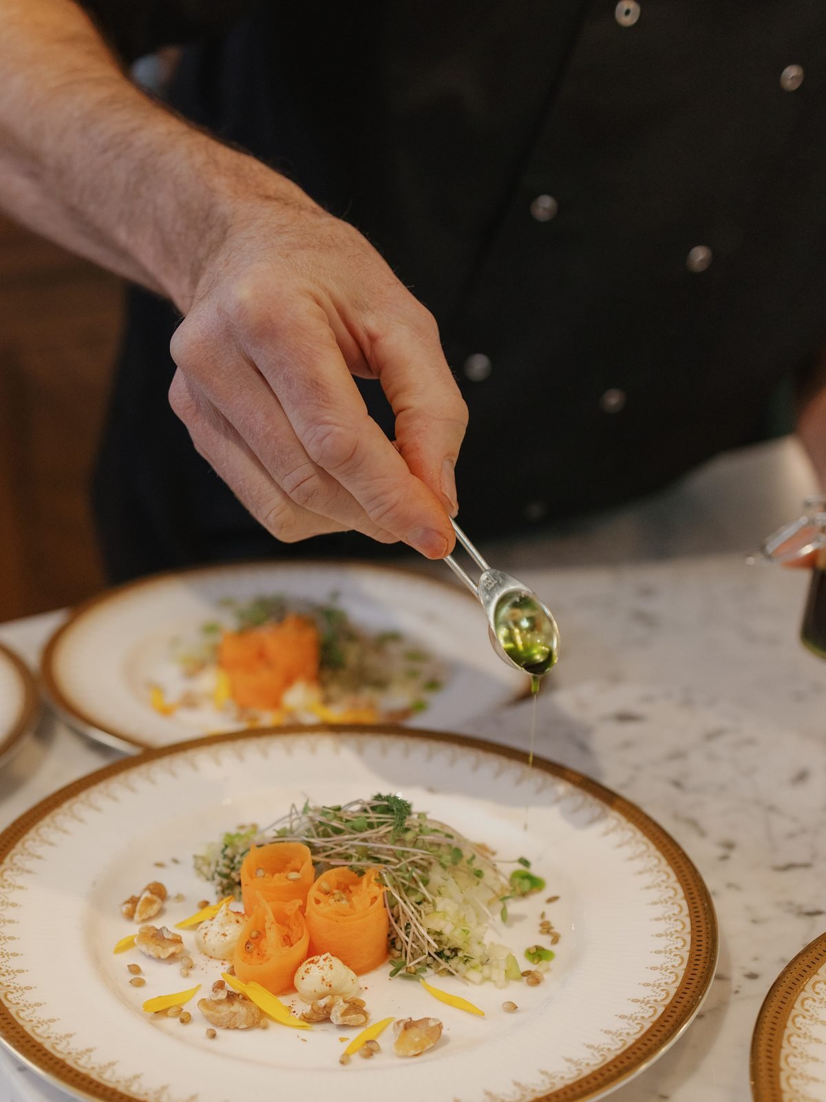 A chef drizzling oil onto a plated dish with thinly sliced carrots, microgreens, and nuts, on a white and gold-rimmed plate.