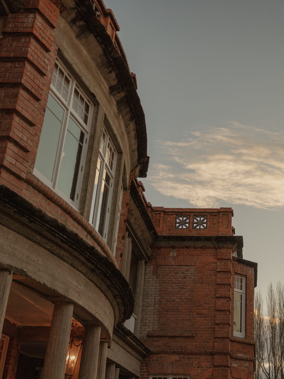 A brick building with large windows and a rounded balcony supported by columns, under a sky with soft clouds at dusk.