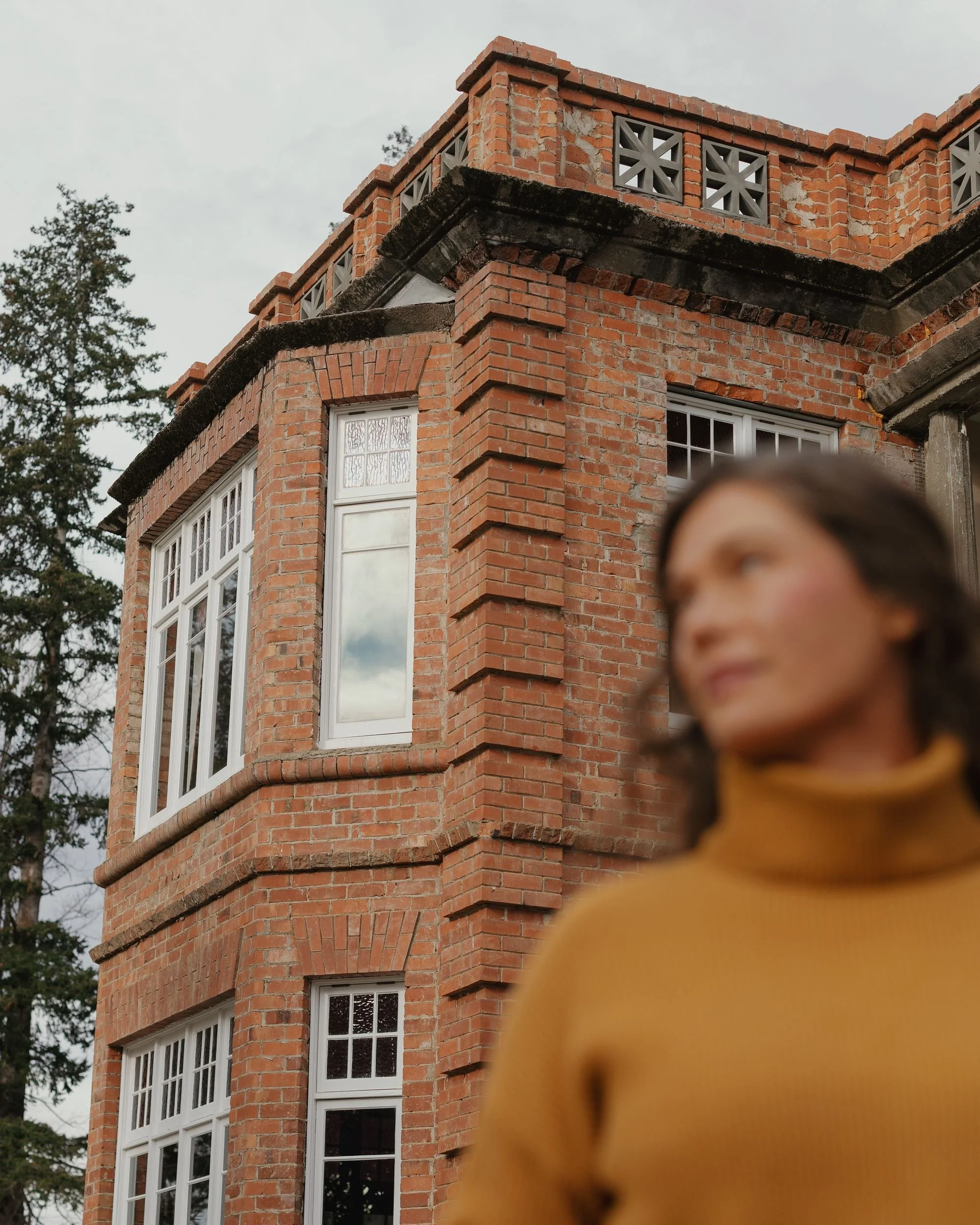 Close-up of a multi-story brick building with white-framed windows and a decorated corner, next to a tall tree, under a cloudy sky. A person in a yellow jacket is partially visible in the foreground.