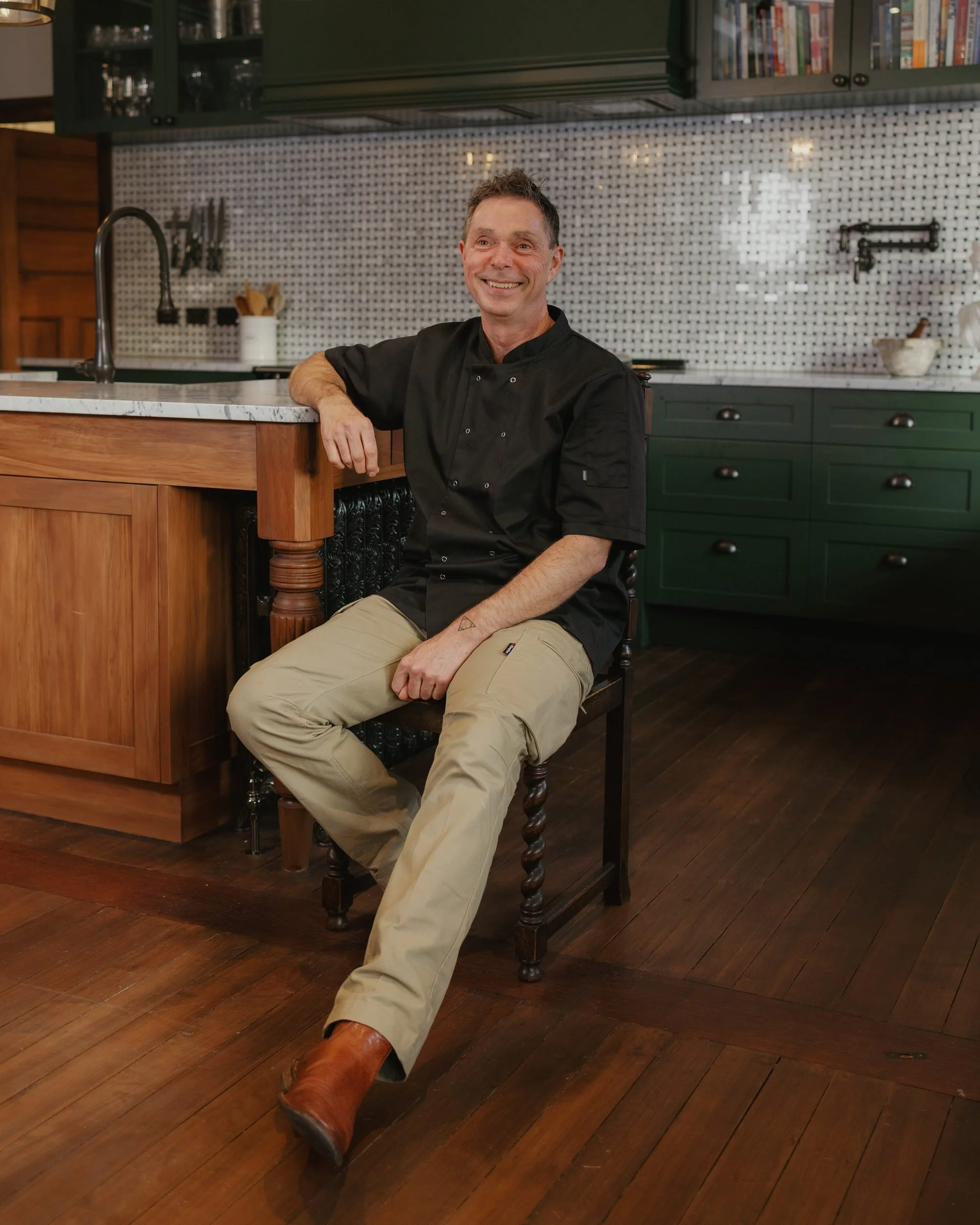 A man in a black chef's coat sitting on a wooden chair in a kitchen with green cabinets and a white tile backsplash, smiling at the camera.