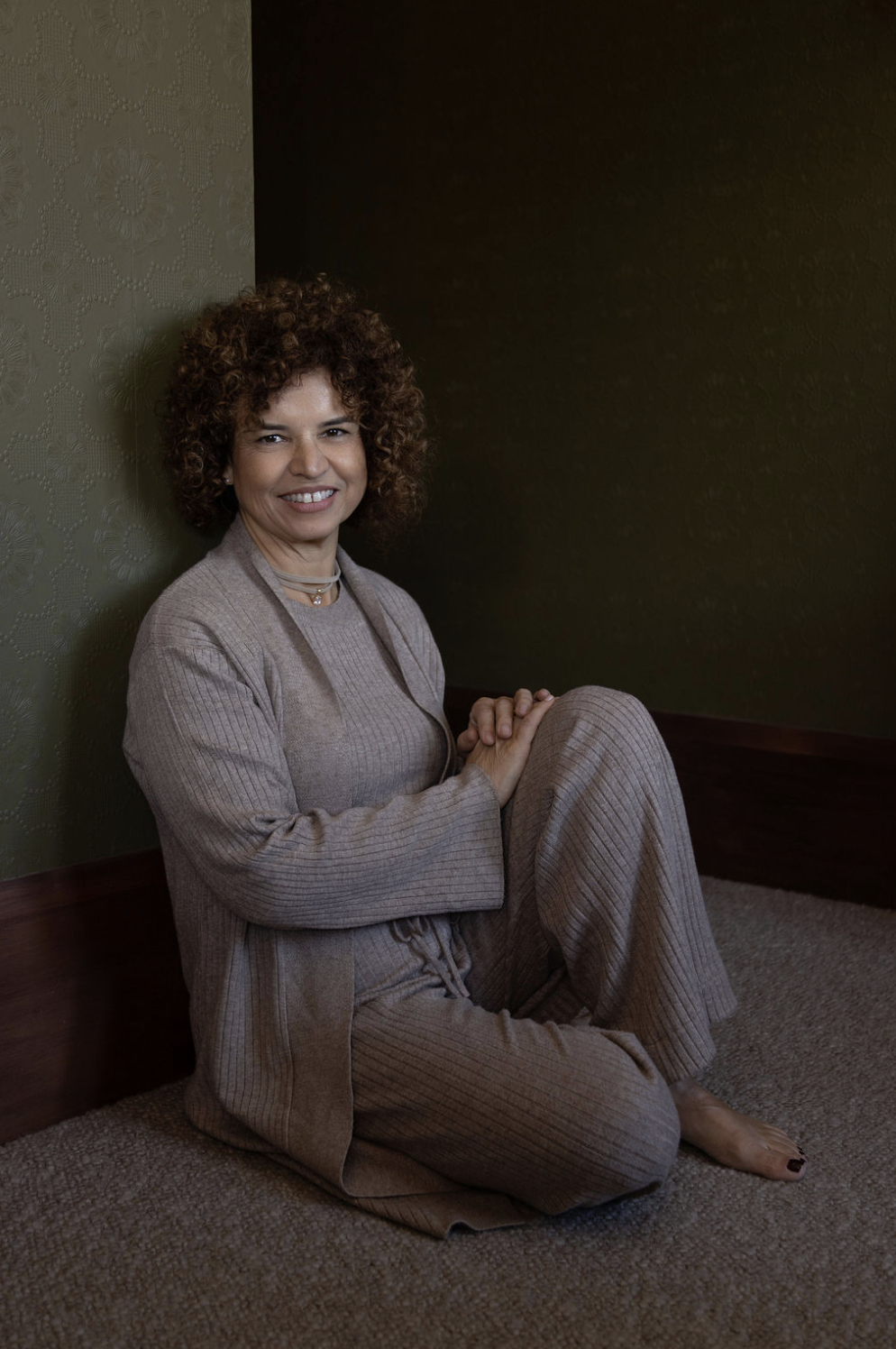 A woman with curly hair smiling, sitting on the floor with her knees bent and hands clasped, wearing beige loungewear, in a room with dark and textured wallpaper.
