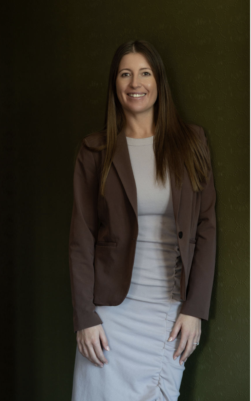 Portrait of a smiling woman with long brown hair, wearing a brown blazer over a light-colored dress, standing against a dark green textured wall.