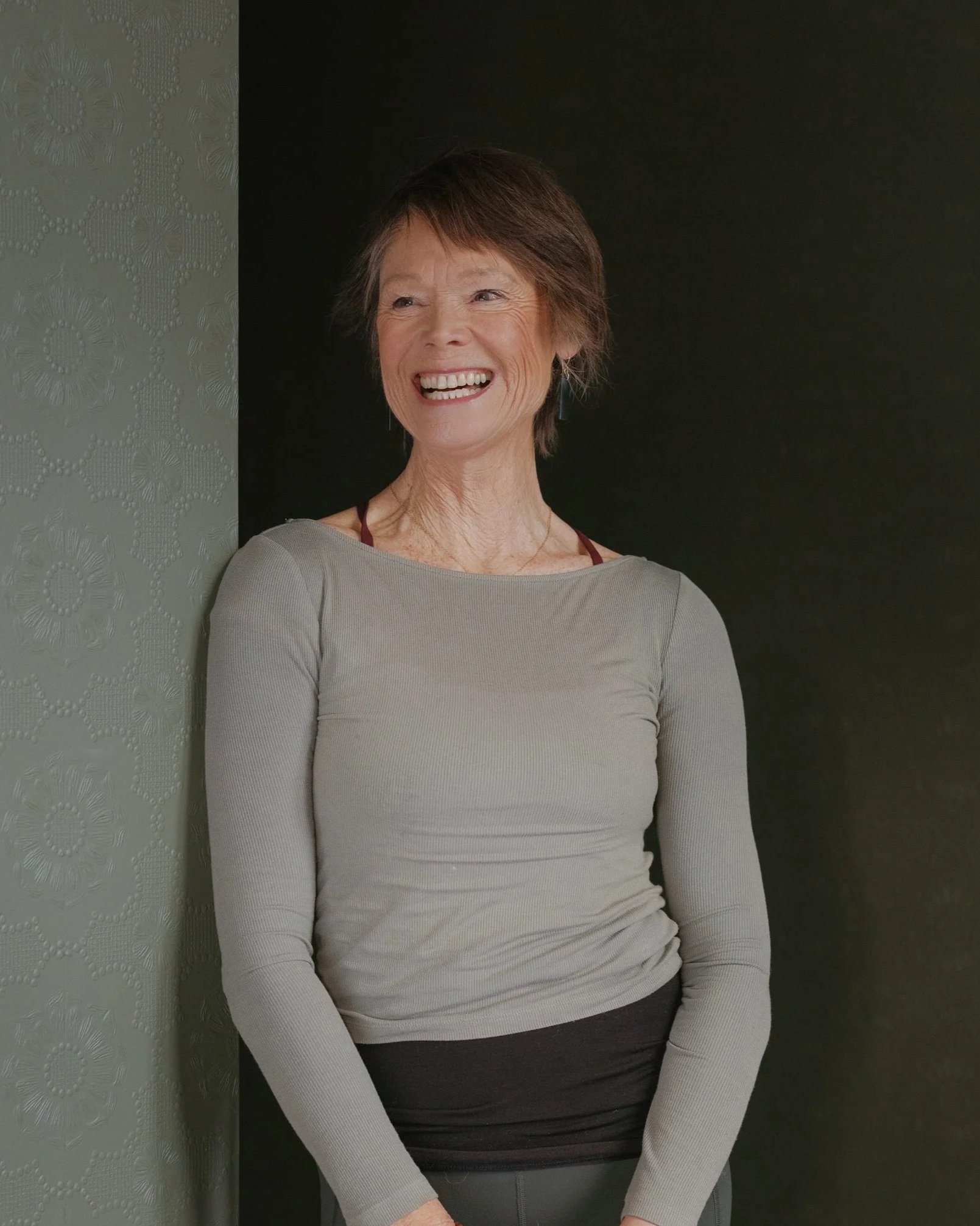 A woman smiling, wearing a light gray long-sleeve shirt and dark pants, standing against a textured gray and black wall.
