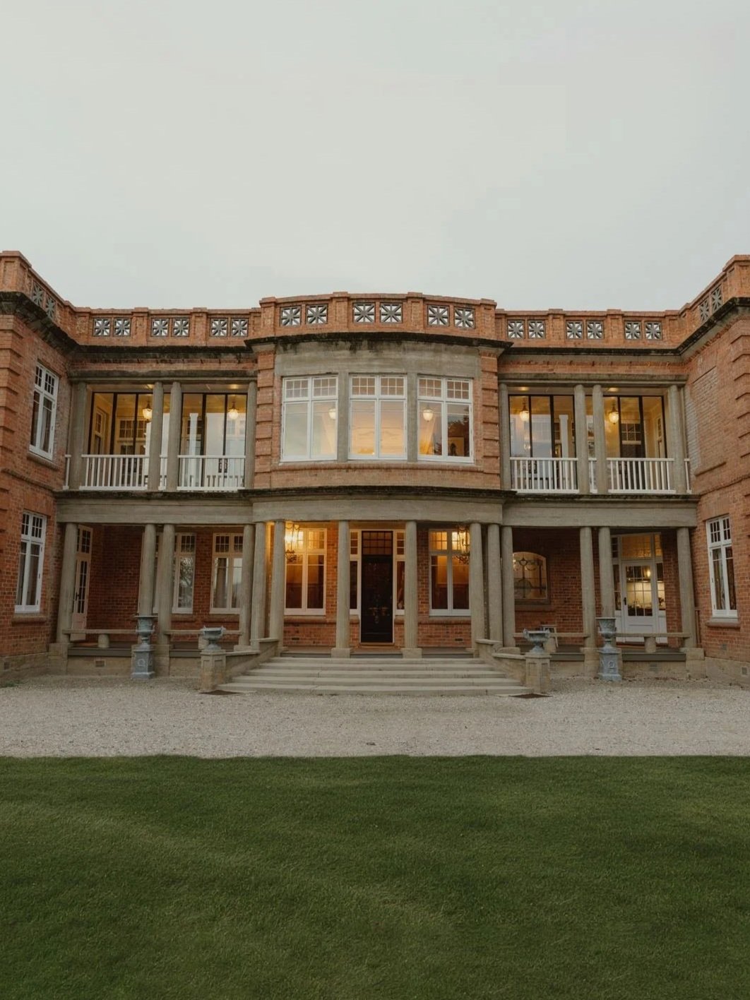 Large brick house with double front stairs, tall columns, and large windows, illuminated from inside, surrounded by a green lawn and gravel pathway.