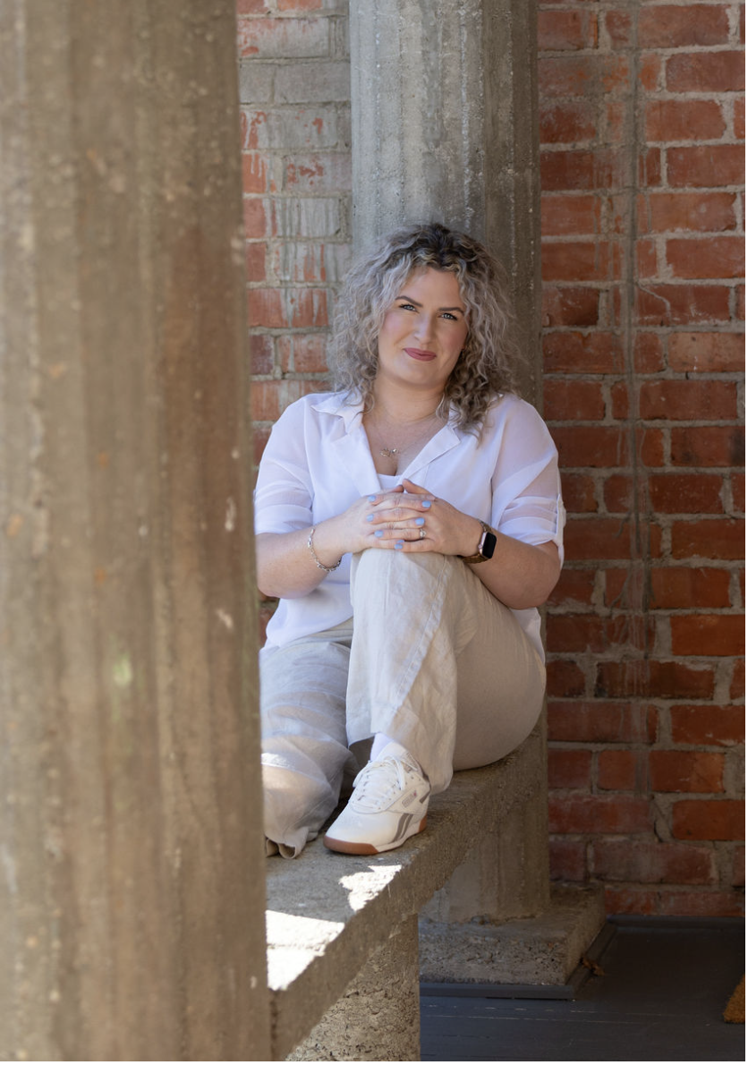 A young woman with curly blonde hair sitting on a concrete ledge between two rough-textured concrete columns in front of a brick wall. She is wearing a white shirt, beige pants, and white sneakers, and is smiling with her hands clasped on her knee.