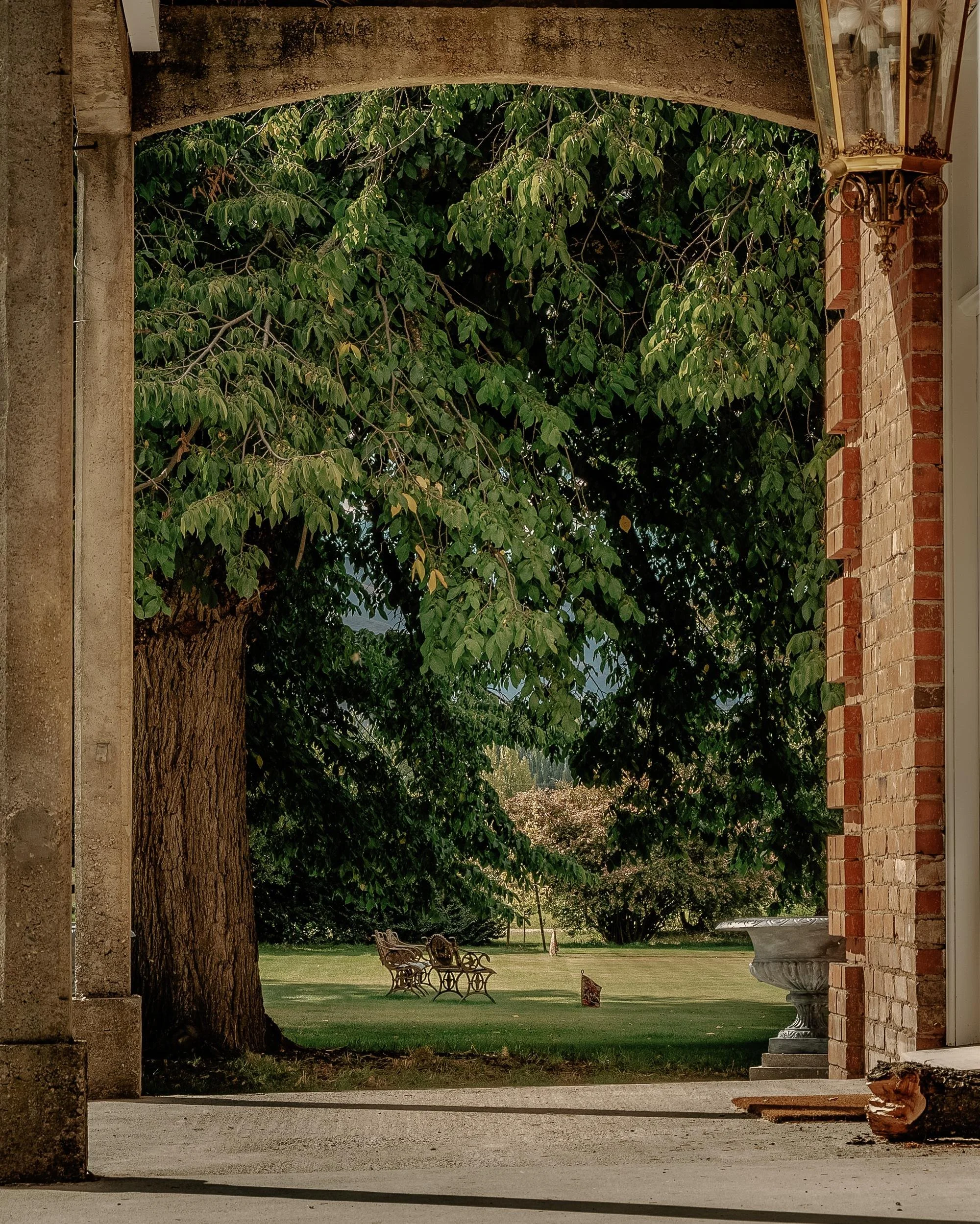 View through a brick archway to a green lawn with two ornate metal benches, a decorative fountain, and trees in the background.