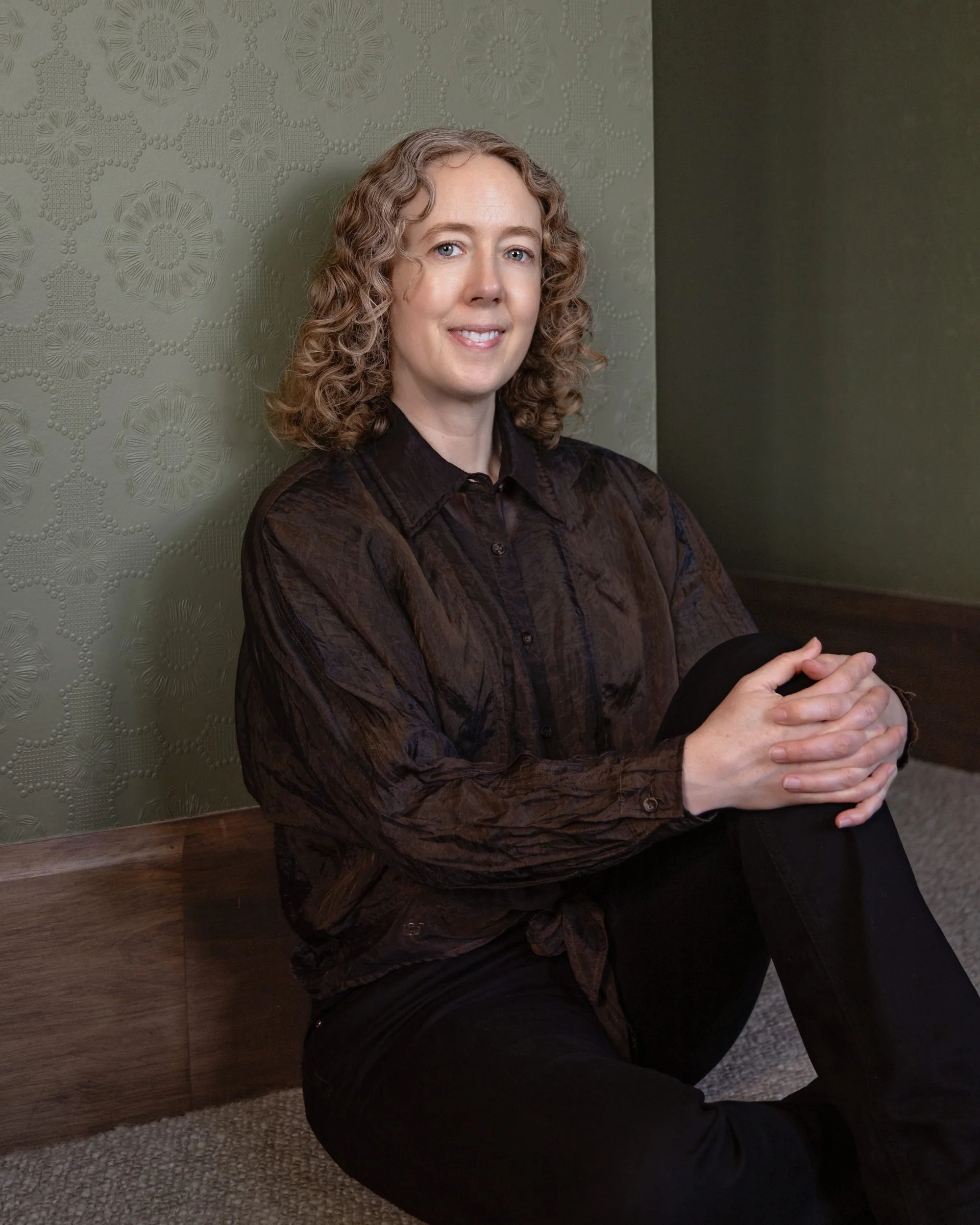 A woman with curly blonde hair and light skin sitting indoors on a wooden ledge, wearing a dark patterned shirt and black pants, with her hands clasped together. The background features green textured wallpaper and a dark wall.