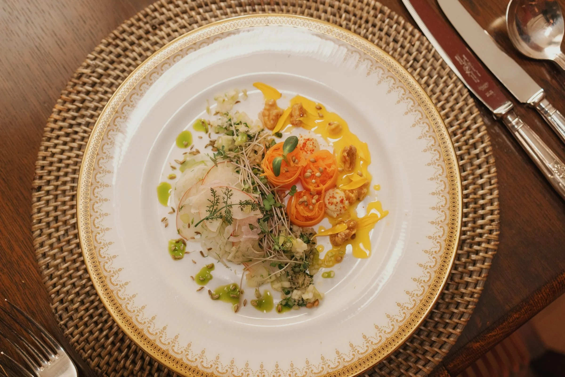 Elegant white plate with gold detailing, holding a colorful assortment of food including garnished vegetables, sauces, and small bites, set on a woven placemat on a wooden table with silverware and a water glass nearby.