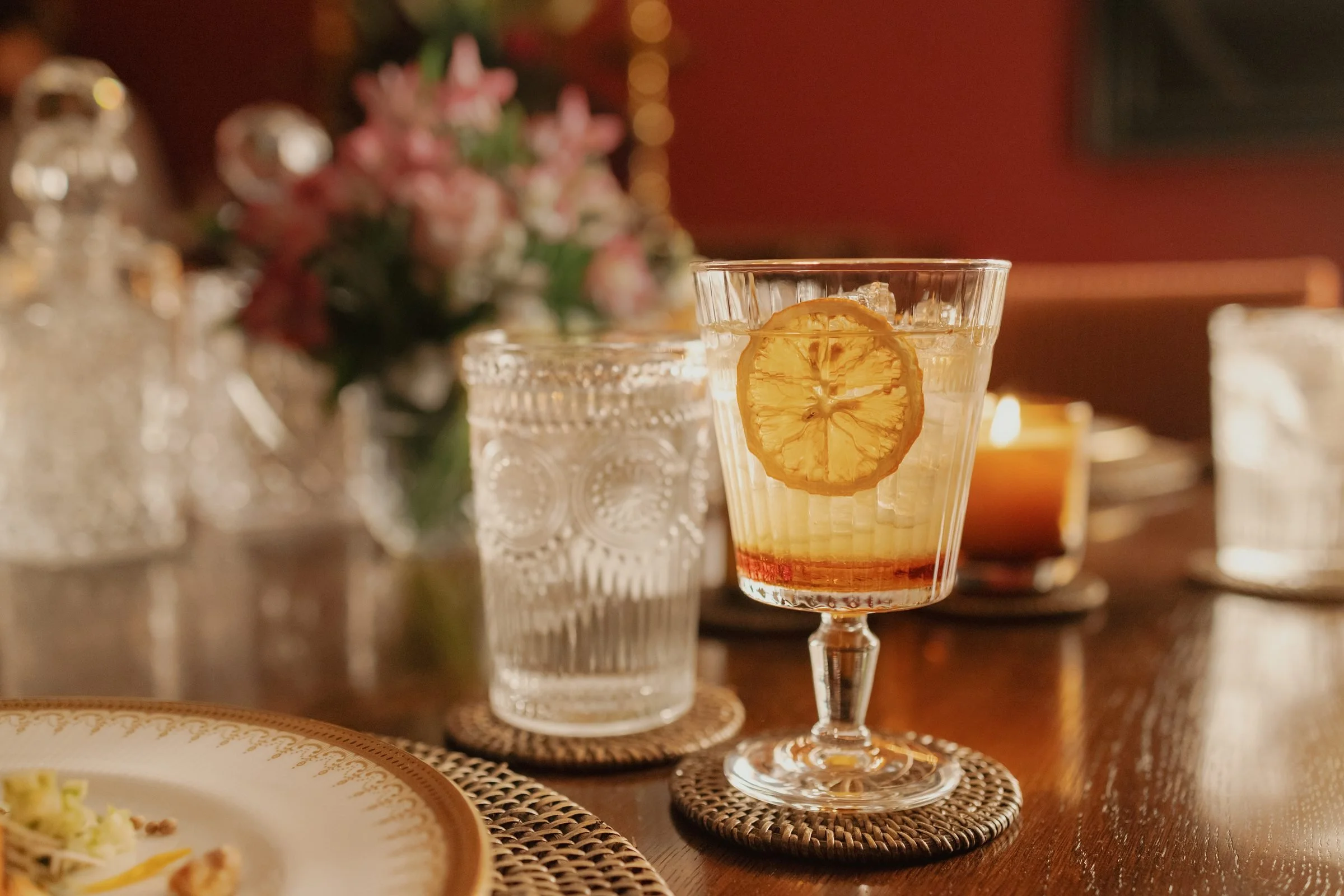A glass with a lemon slice in it sits on a wine coaster on a wooden table, with blurred decorative objects and flowers in the background.