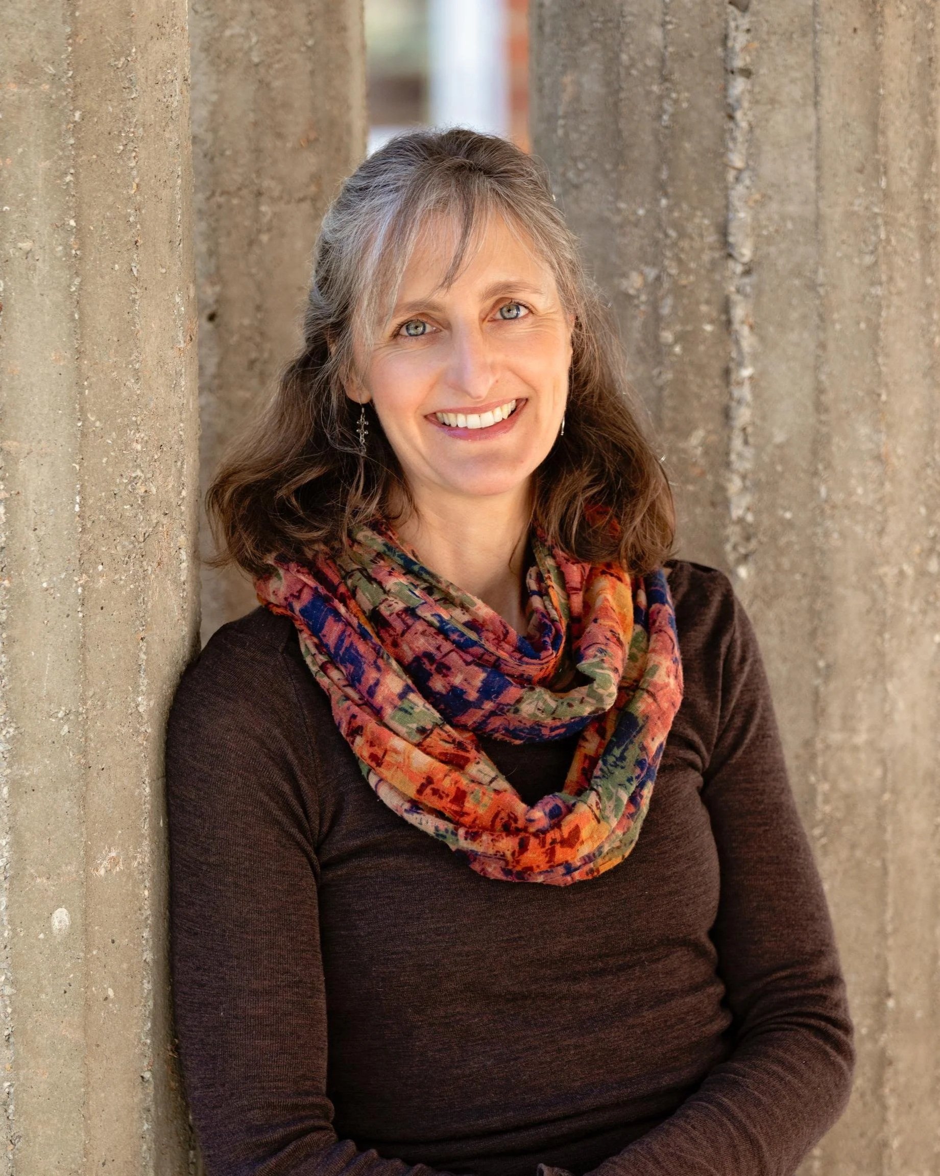 A woman with gray hair, blue eyes, and earrings, smiling, wearing a colorful scarf and a dark top, standing next to a concrete wall.