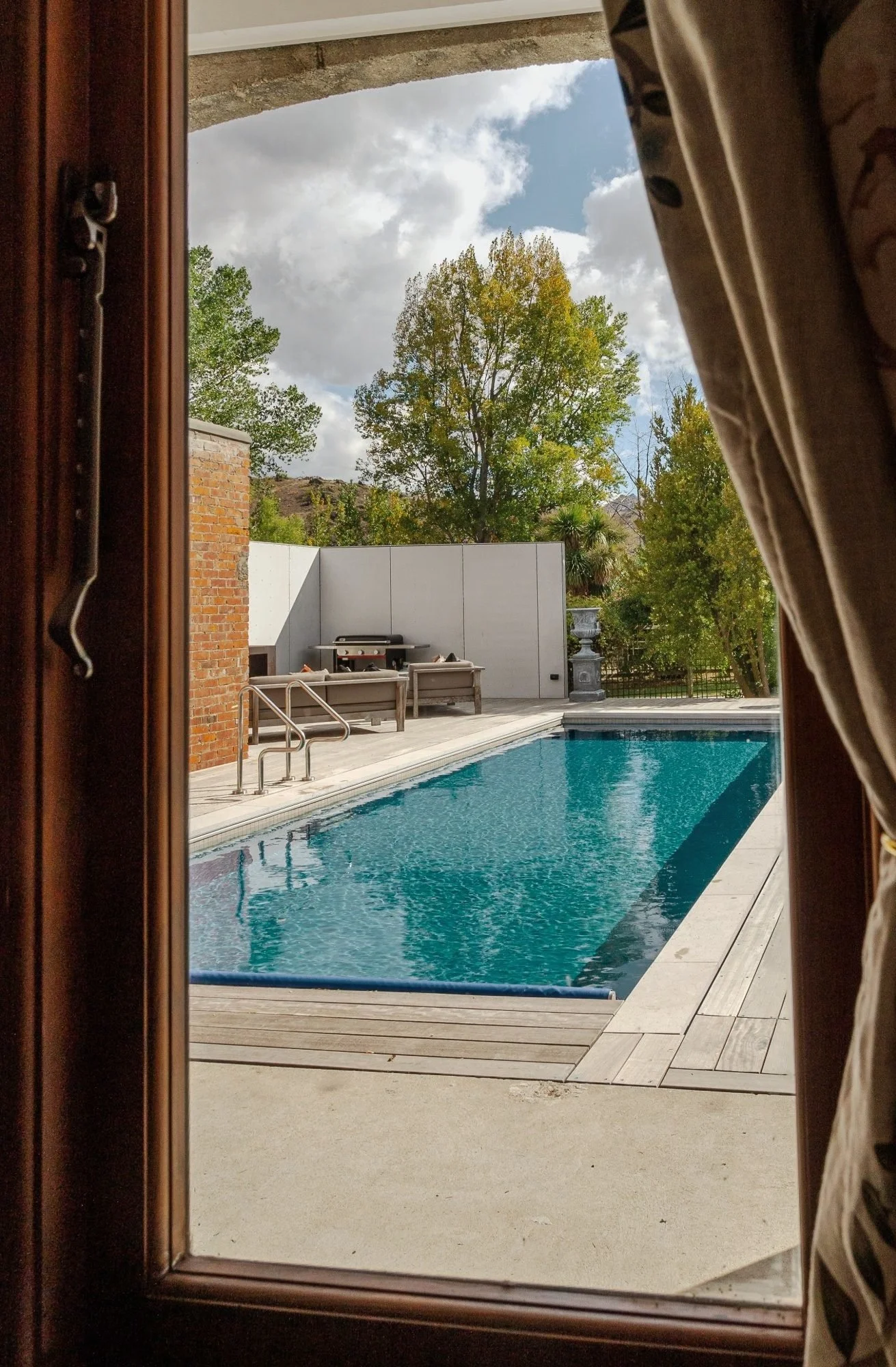 View through a glass door showing an outdoor swimming pool, poolside benches, a barbecue grill, and a white fence with trees and a cloudy sky in the background.