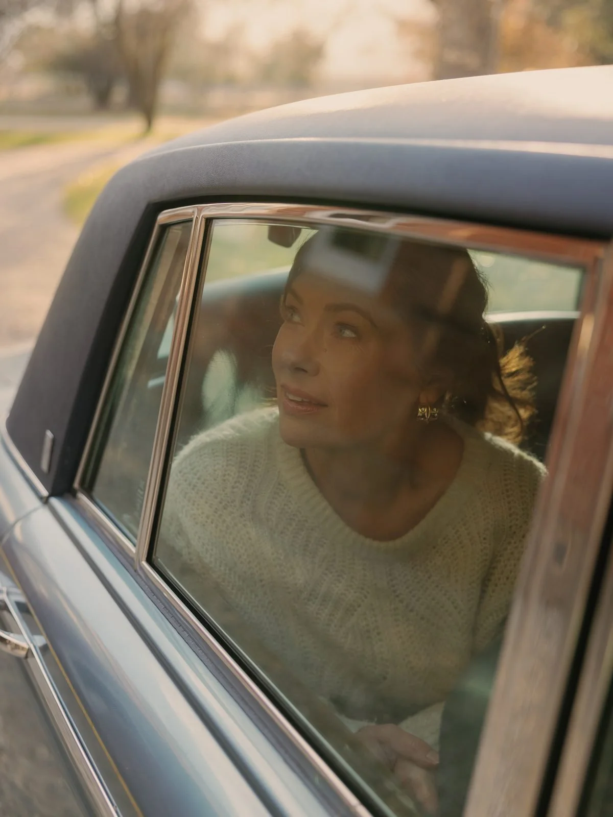 A woman with brown hair and gold earrings looks through the side window of a vintage car, with a soft focus on a rural landscape in the background during sunset.