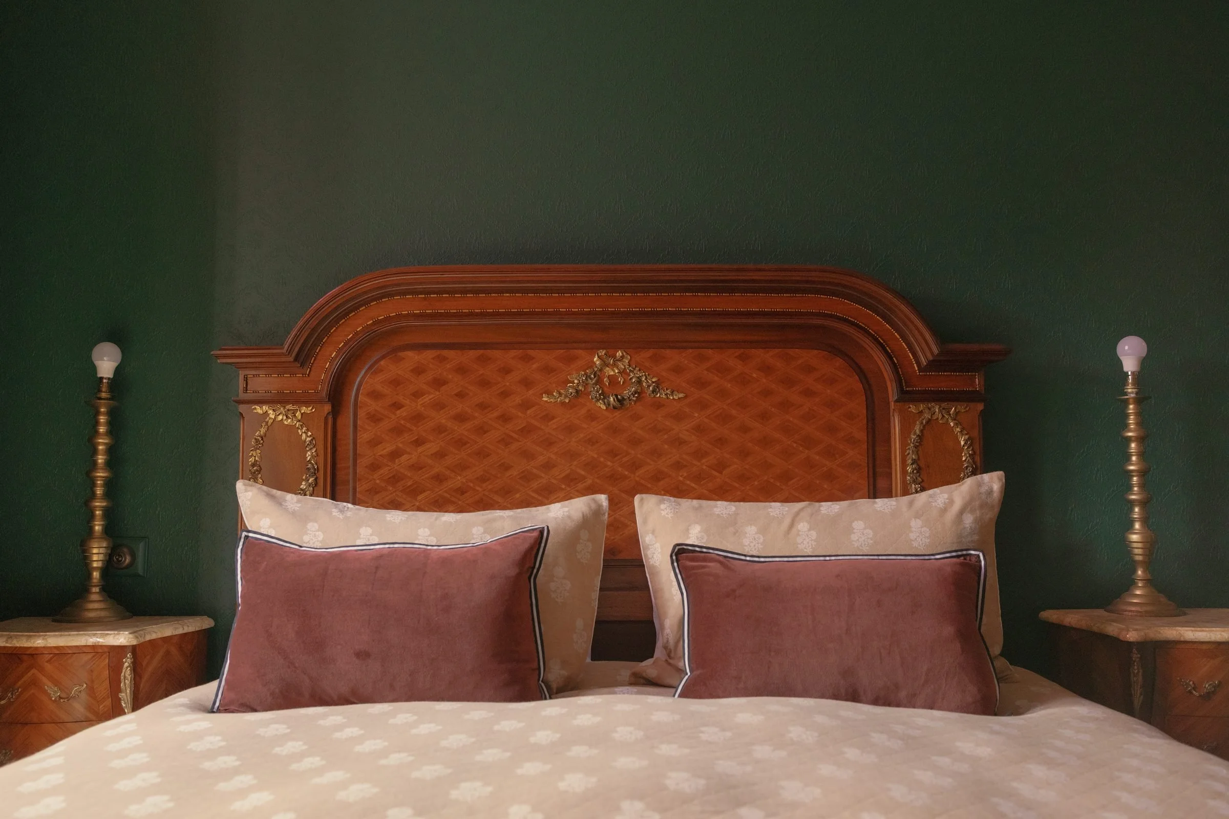 A bedroom with a green wall, a wooden headboard with gold accents, two lamps with brass bases, two pillows with beige pillowcases and decorative pillows with brown and beige fabrics, and small wooden side tables.