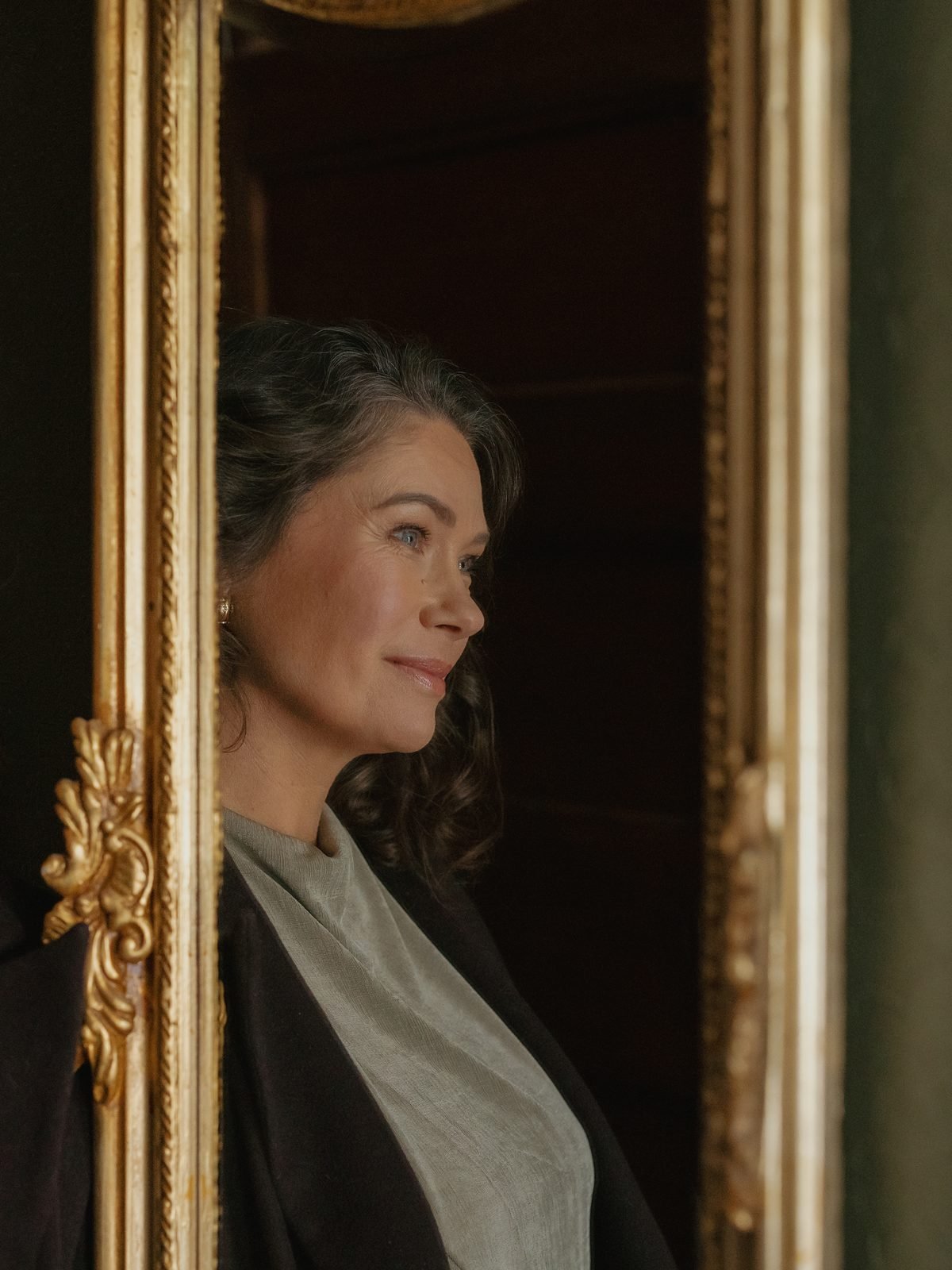 A woman with gray hair and a gray scarf looking to the left, seen through a decorative gold-framed mirror.