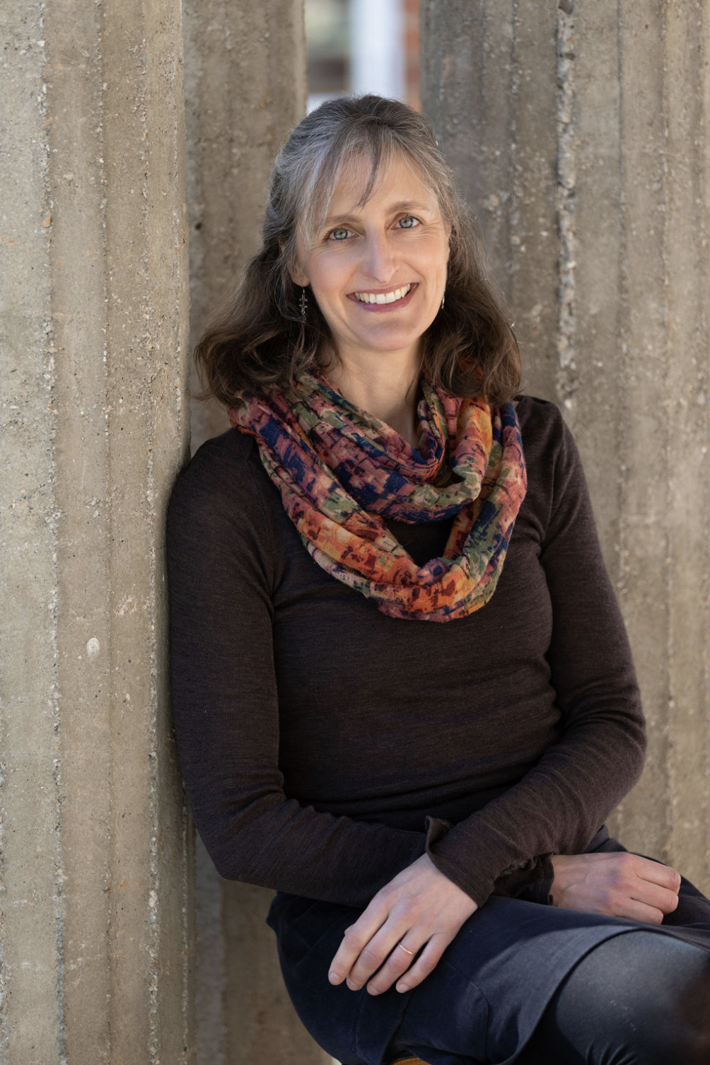 A woman with shoulder-length gray hair smiling, wearing a black shirt, colorful scarf, and dark pants, leaning against concrete pillars outdoors.