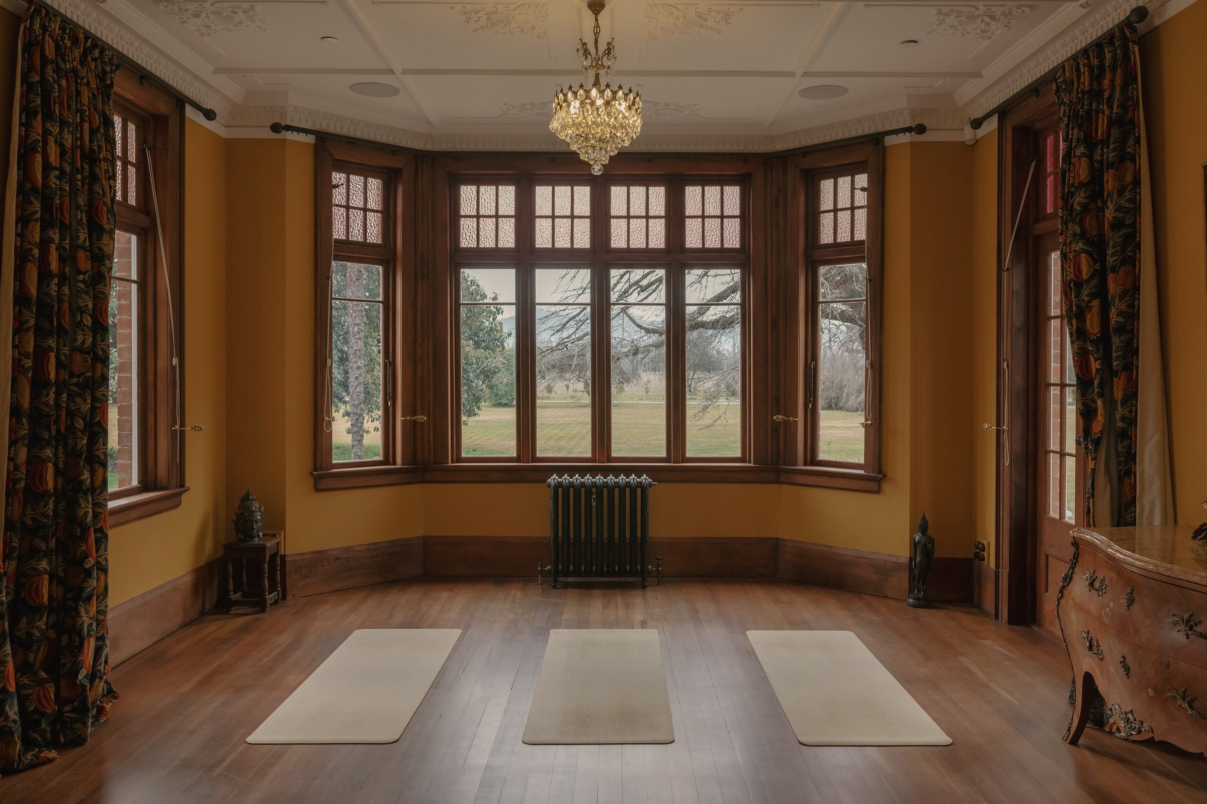 A spacious living room with large windows and a view of trees outside, decorated with a chandelier, patterned curtains, and wooden furniture.