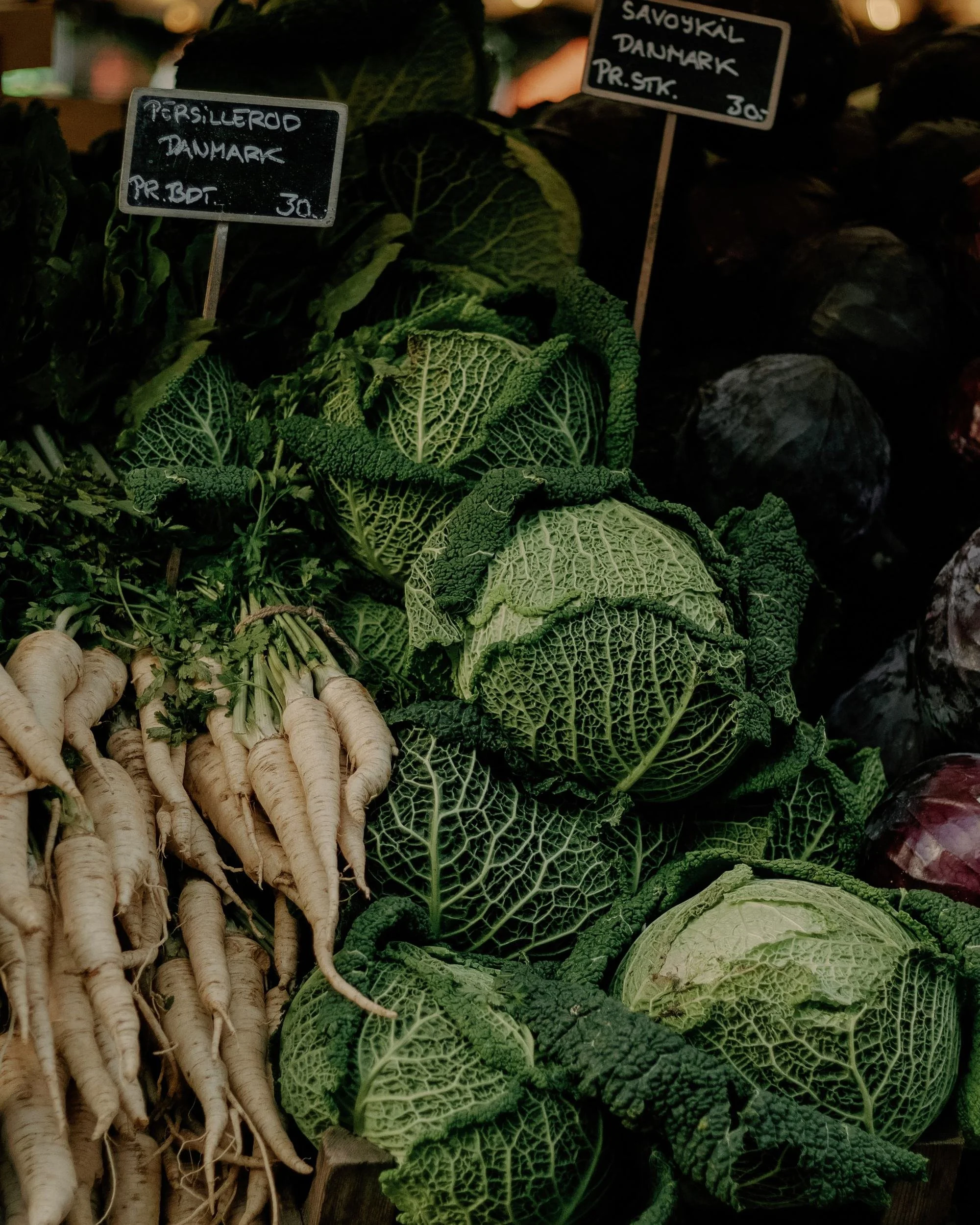 Fresh vegetables including white parsnips, green savoy cabbage, and other leafy greens displayed at a market stall, with handwritten price signs in Danish kroner.