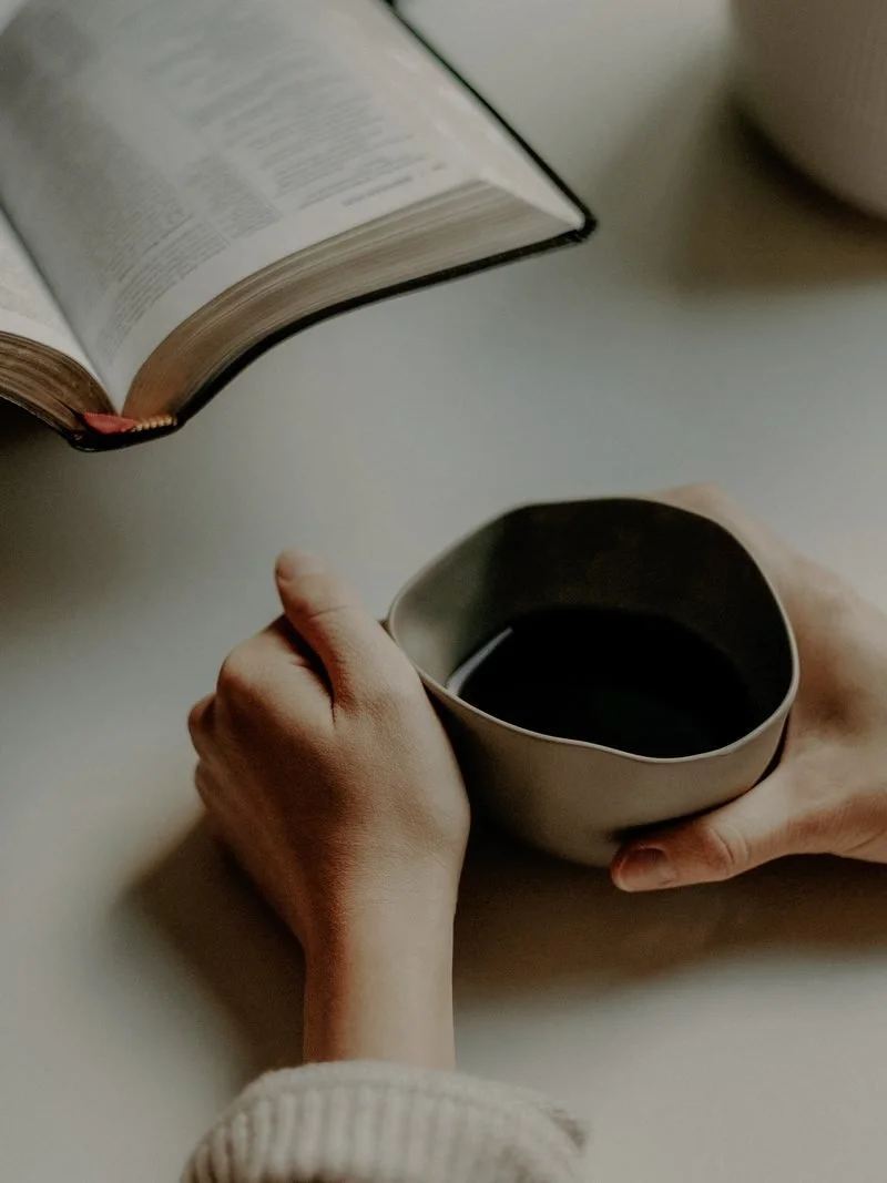 Person holding a mug of dark coffee next to an open book on a white table.