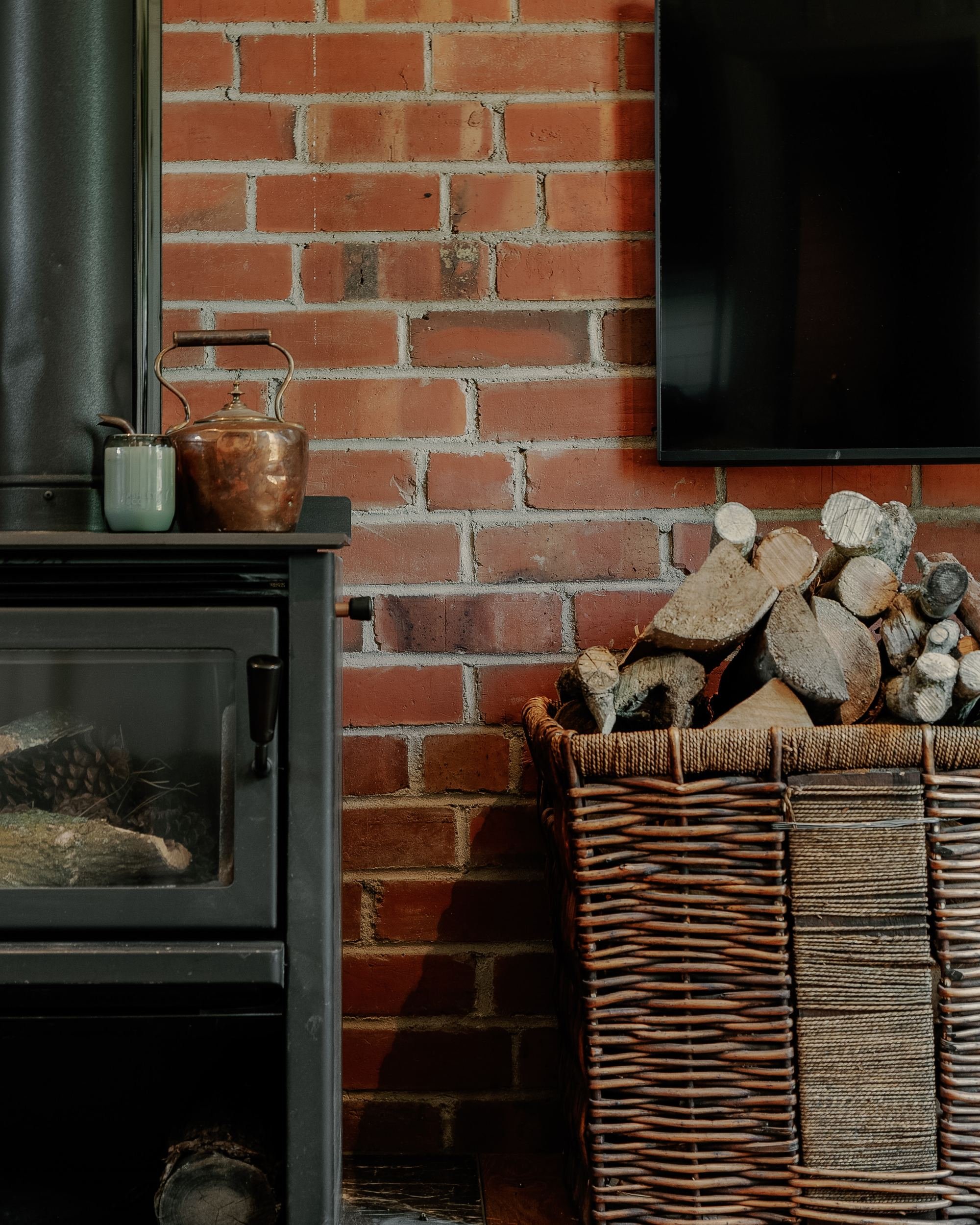 A brick fireplace with a wood stack, a wicker basket, a television on the wall, and a small stove with decorative pots on top, set against a red brick wall.