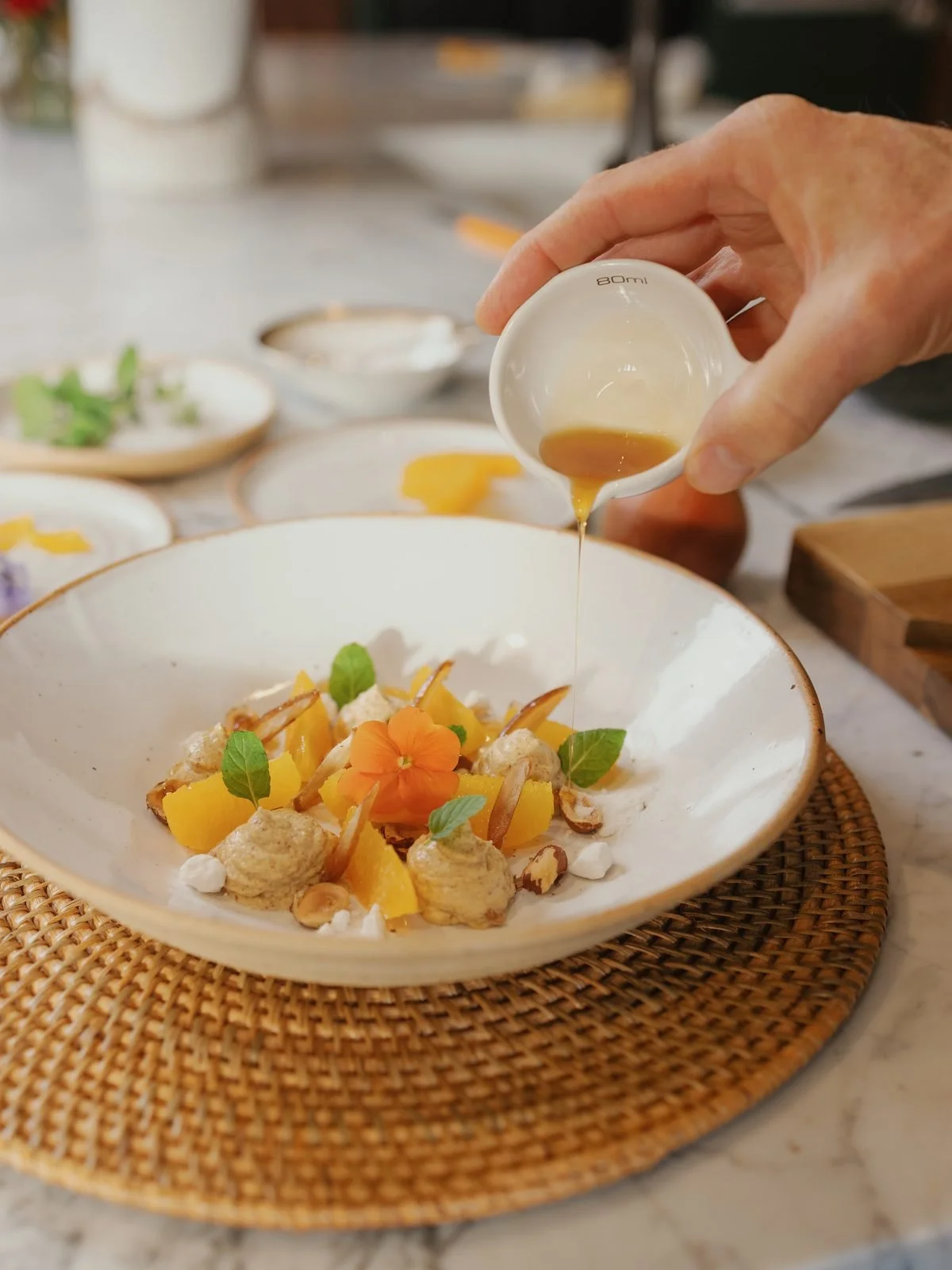A person pouring a golden sauce onto a plated dish with flowers and green leaves, set on a woven mat.