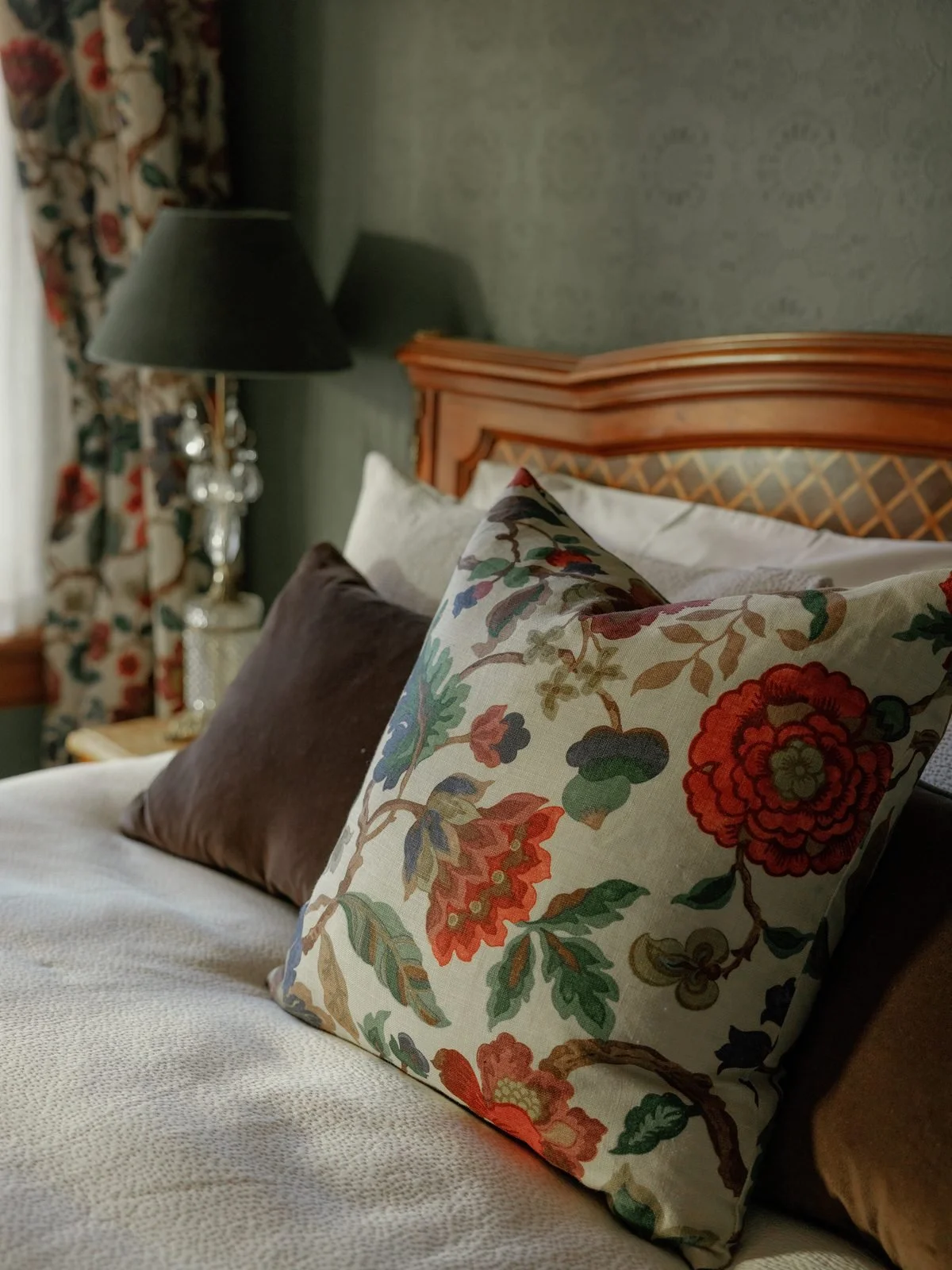 A bedroom with floral and solid pillows on a bed, a wooden headboard, a black lampshade, a decorative lamp, patterned curtains, and textured walls.