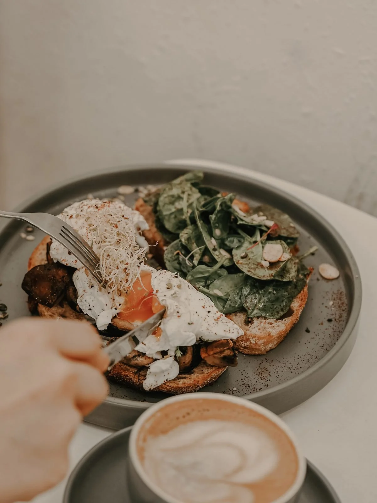 Close-up of a plate with two slices of toasted bread topped with spinach and an egg, with shredded cheese and sautéed mushrooms, next to a cup of frothy coffee.