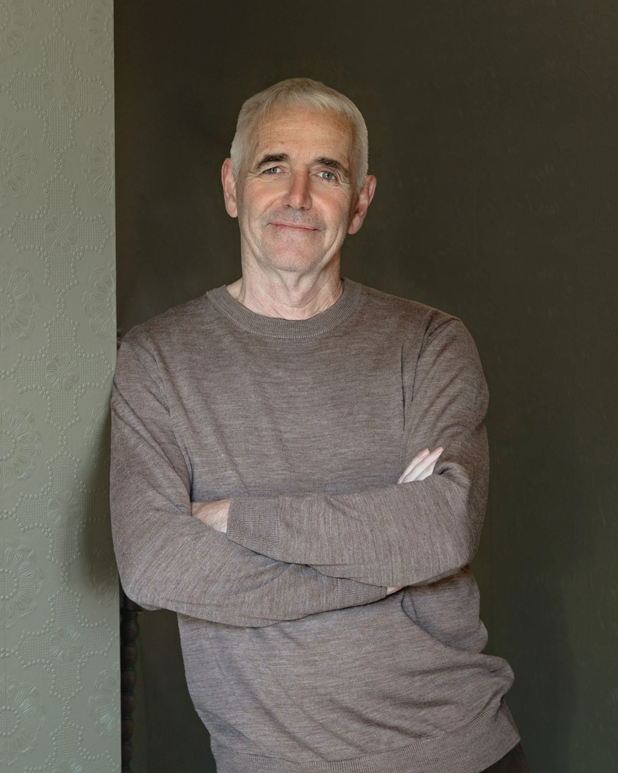 A middle-aged man with gray hair and blue eyes, standing with arms crossed, smiling softly, in front of a dark wall with textured wallpaper on the left side.