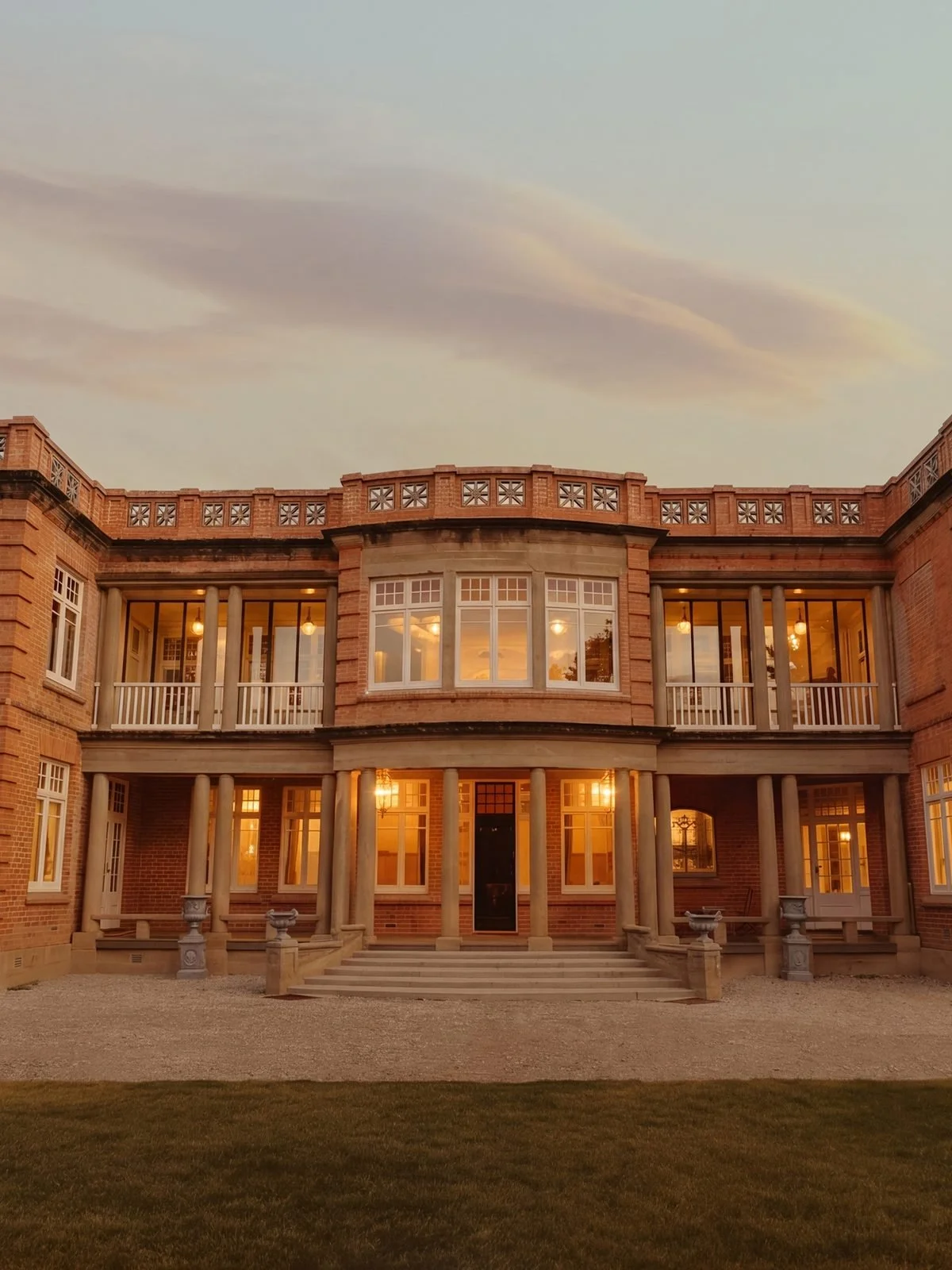 A large brick mansion with columns, multiple windows, and a balcony, illuminated with warm indoor lighting during sunset.