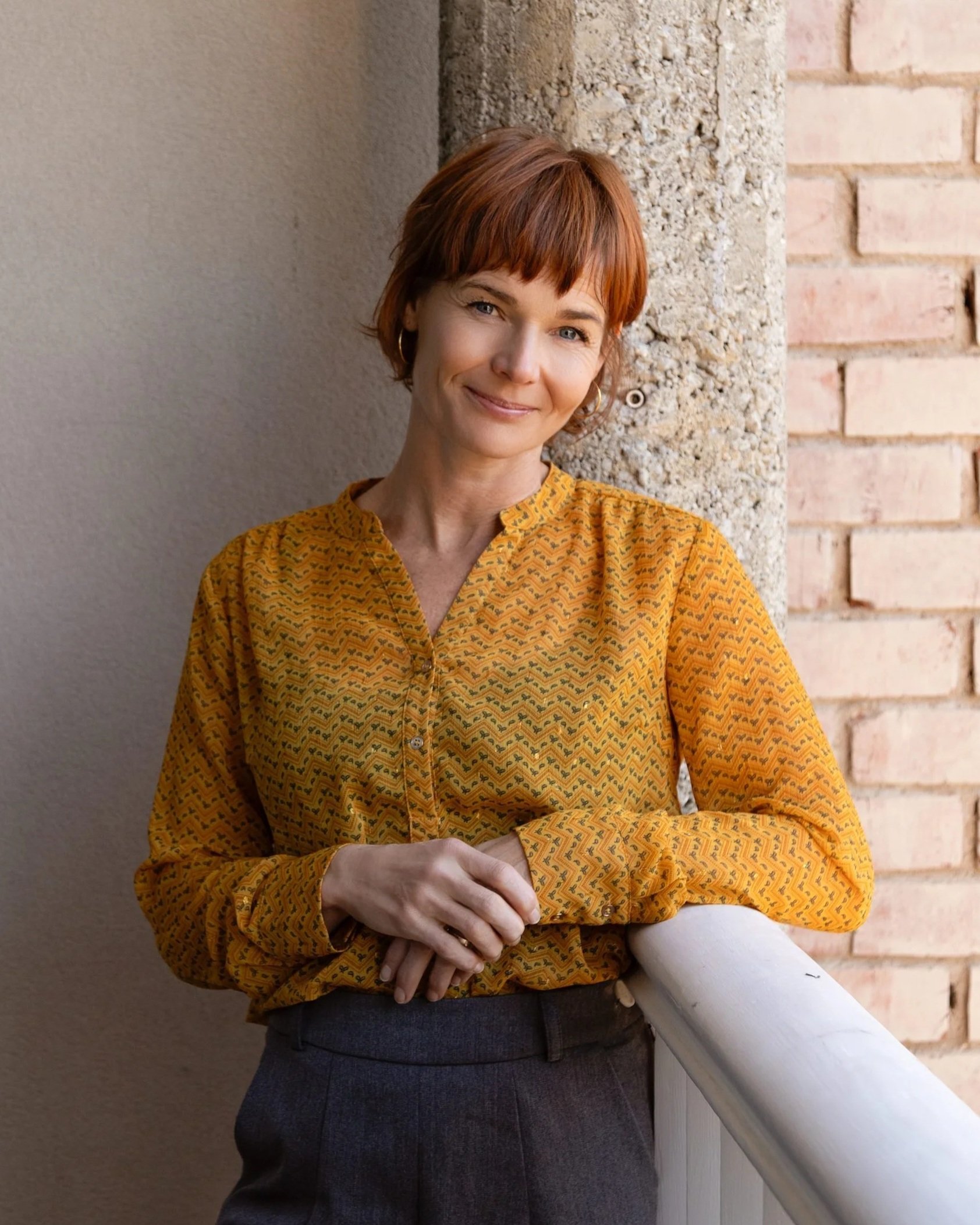 A woman with short red hair wearing a yellow patterned blouse and dark pants, standing outdoors near a brick wall and holding a rolled-up paper.