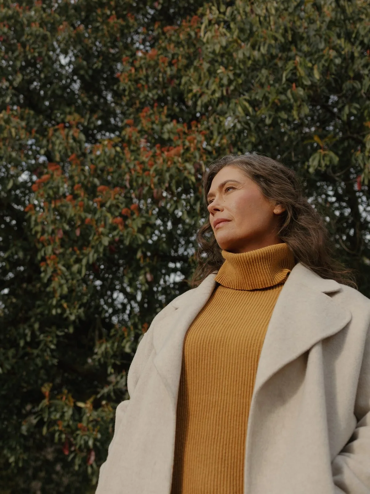 A woman with brown, curly hair standing outdoors in front of a tree with green and orange leaves, wearing a beige coat over a mustard yellow turtleneck sweater, looking slightly upward with a calm expression.