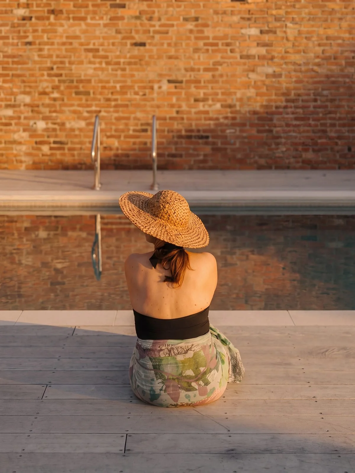 A woman, sitting by a pool, wearing a sun hat and a floral sarong, facing away from the camera towards a brick wall.