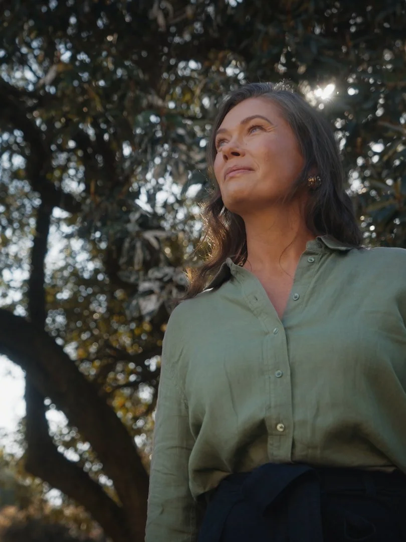 A woman with dark hair, wearing a green button-up shirt, standing outdoors in front of a tree with sunlight filtering through the leaves.